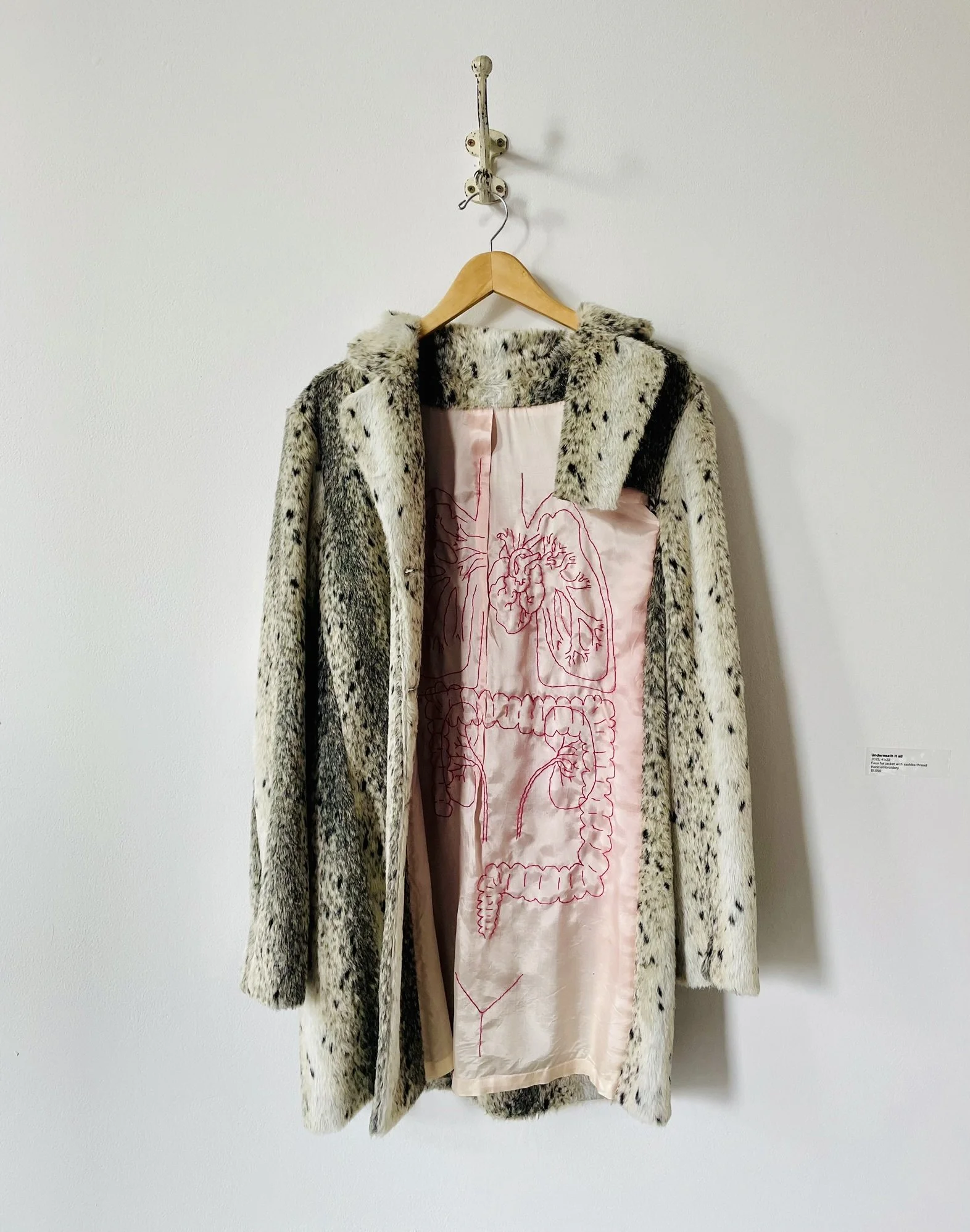 A faux fur coat hung on a hanger with a pink fabric interior featuring embroidered outlines of the human heart and digestive system.