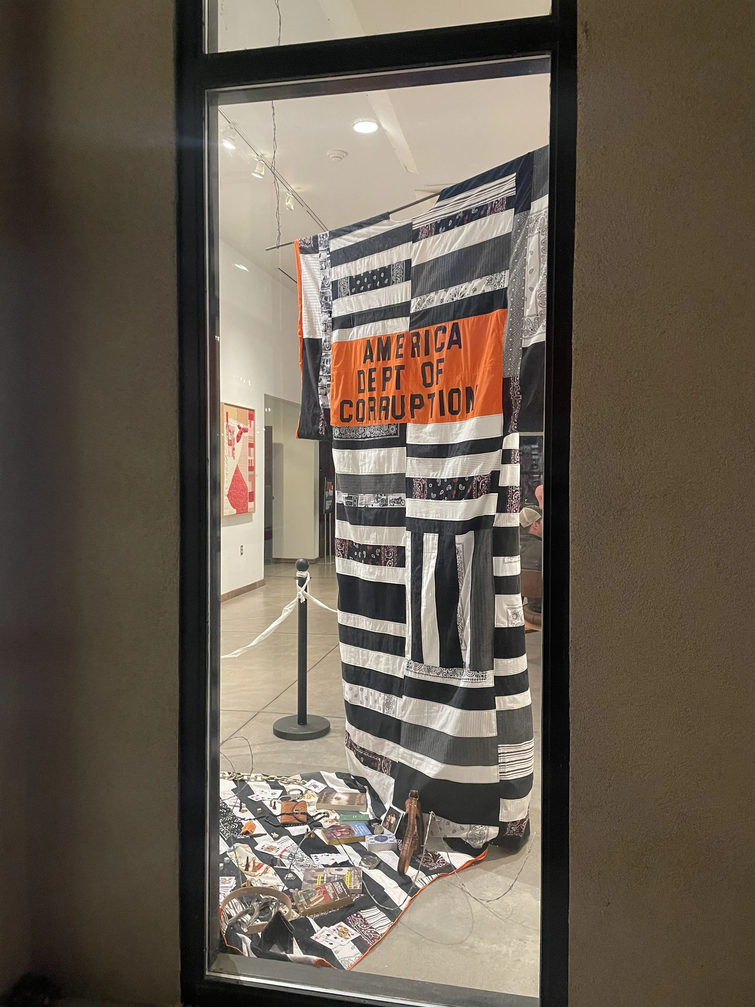 A large window in an art gallery featuring a black and white striped cloth with an orange section that reads 'America Dept of Corruption,' hanging inside a glass showcase.