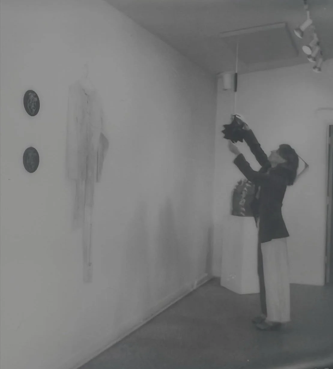 A person in a black jacket and white pants is cleaning or dusting a white wall with a cloth in a room, while standing on the floor; the room has some wall decorations and a ceiling-mounted light fixture.