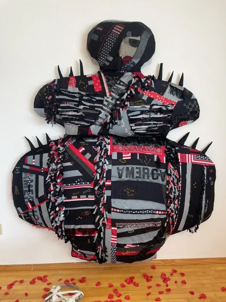 A wall art piece shaped like a person wearing a hoodie and jacket, made from cut fabric with patches of American flag and red, black, gray, and white patterns. Red rose petals are scattered on the floor below.