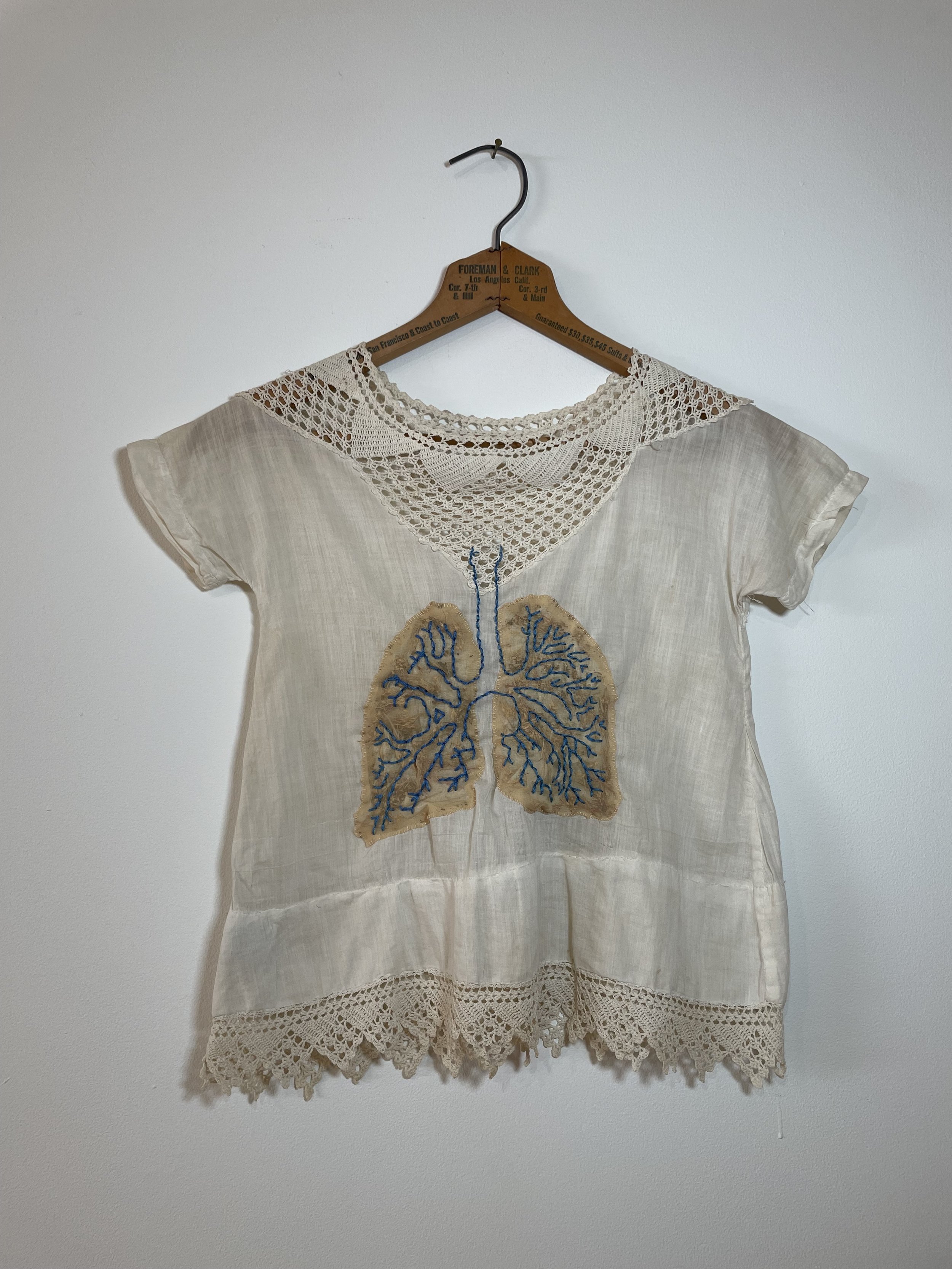 Cream-colored linen shirt with lace crochet neckline and hem, featuring embroidered lungs with blue and tan threads.