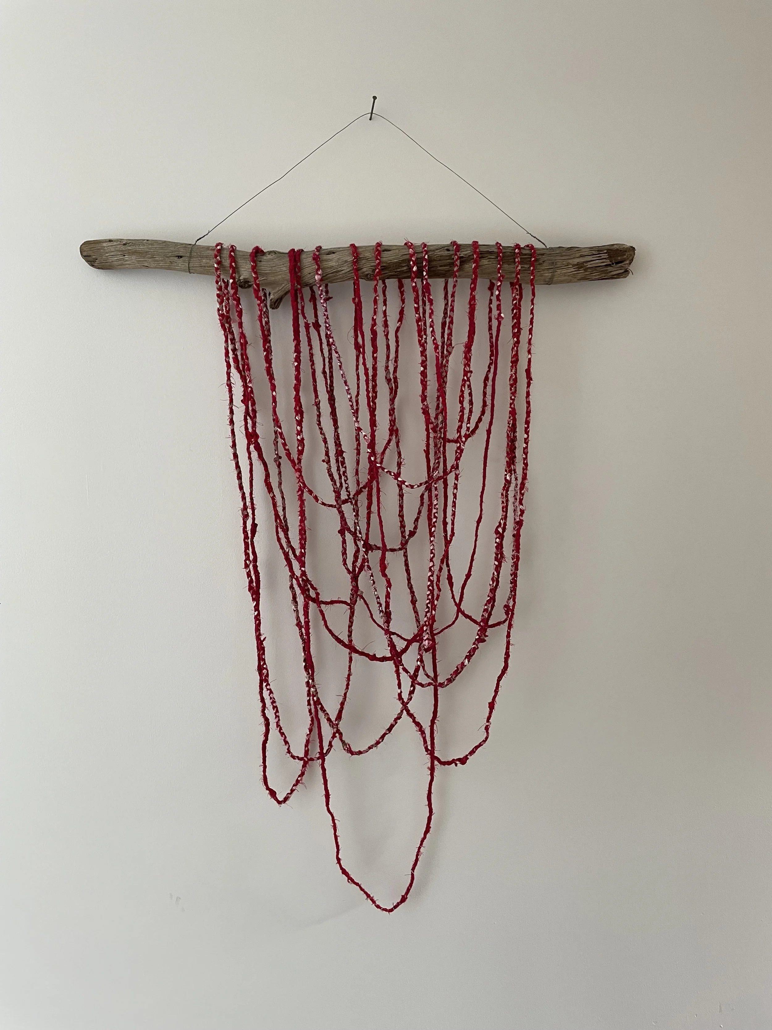 A decorative piece made of a driftwood branch hanging on a white wall, with multiple red braided repurposed textiles draped from it.