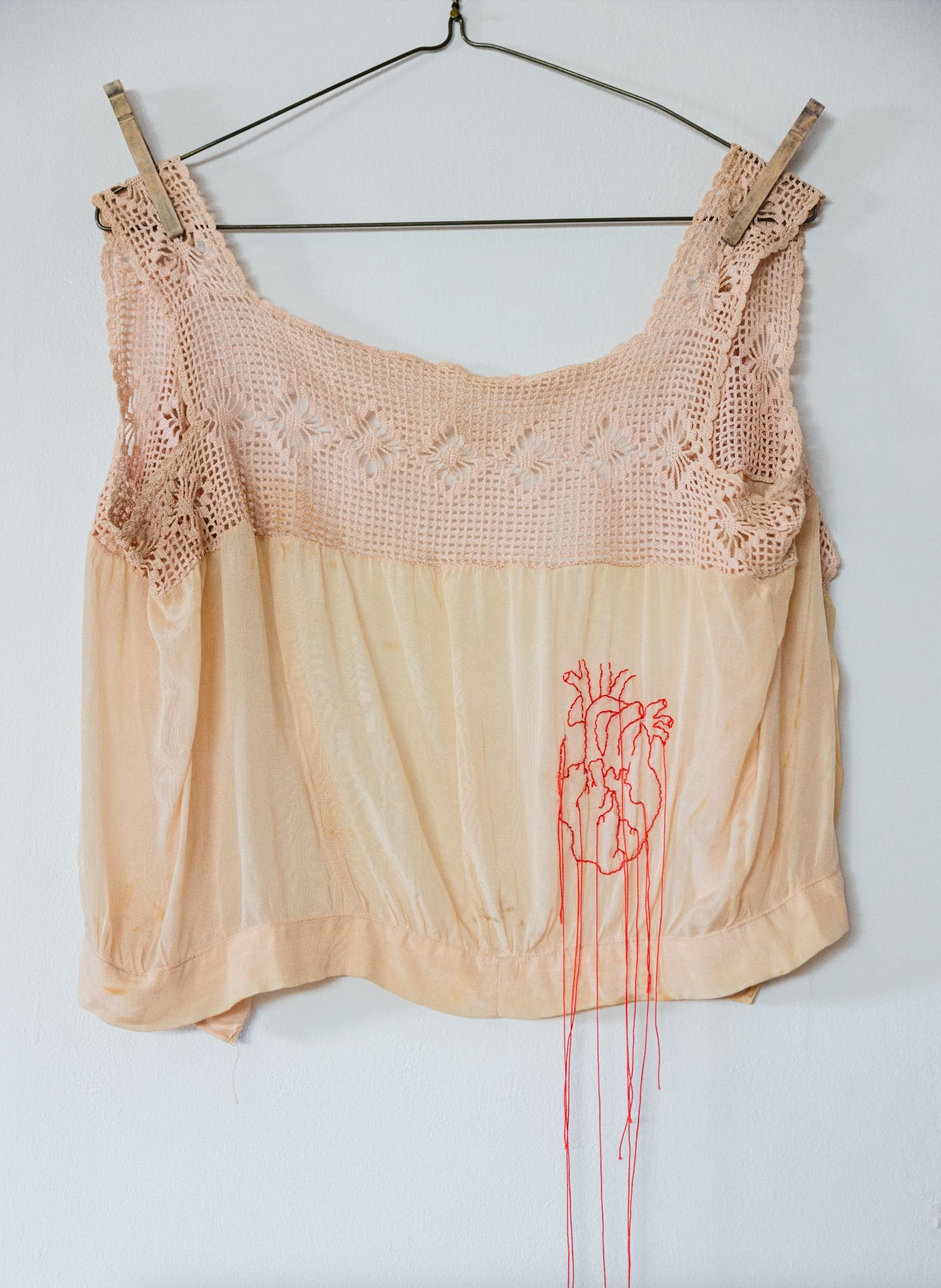 A vintage turn-of-the-century crochet lace top with detail at the neckline, hanging on a wire hanger with clothespins, featuring a red embroidered anatomical heart with hanging threads design.