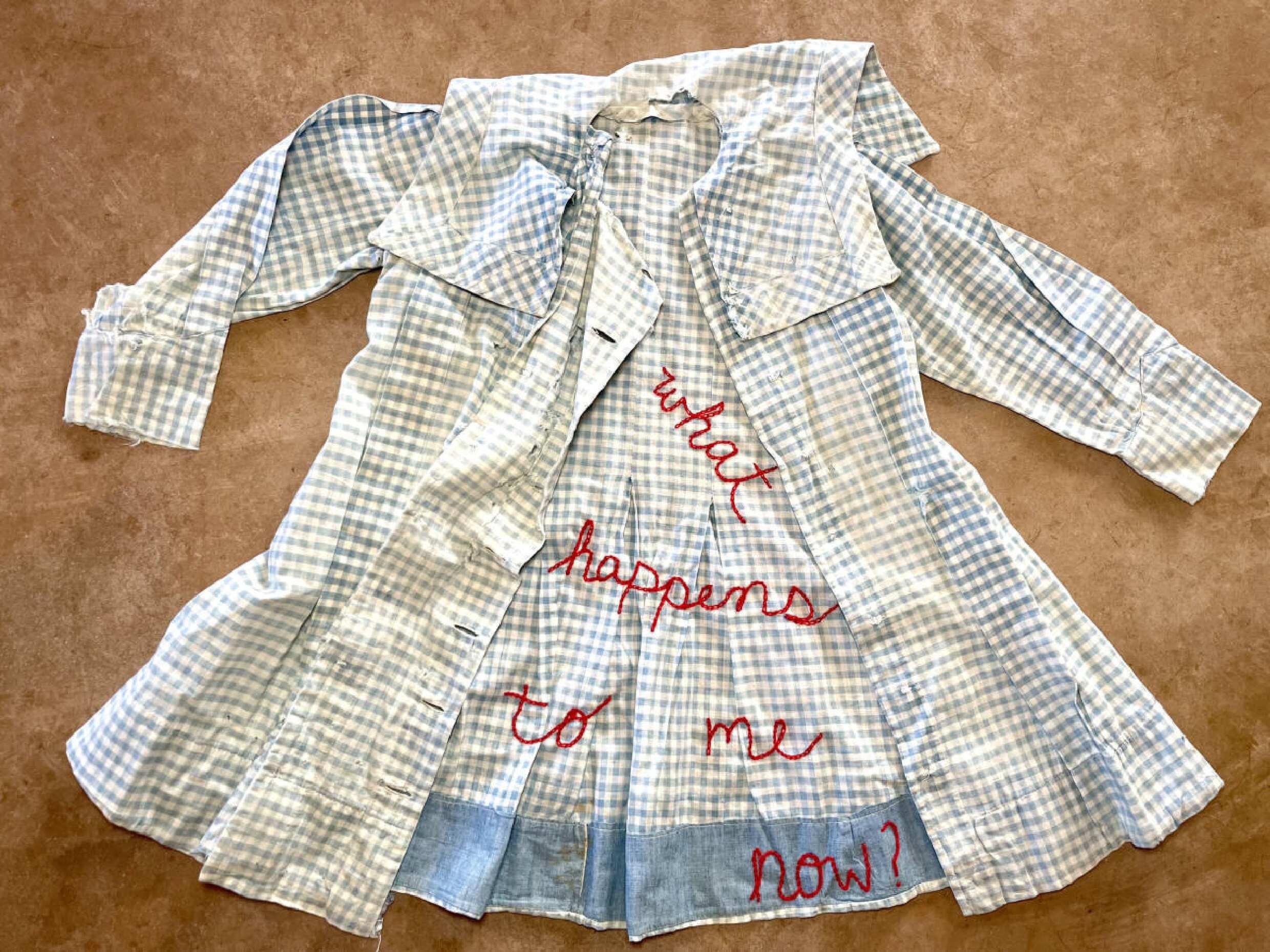 A torn, sleeveless, and damaged blue checkered shirt laid flat on a brown surface. Red embroidered text on the shirt reads "what happens to me now?".
