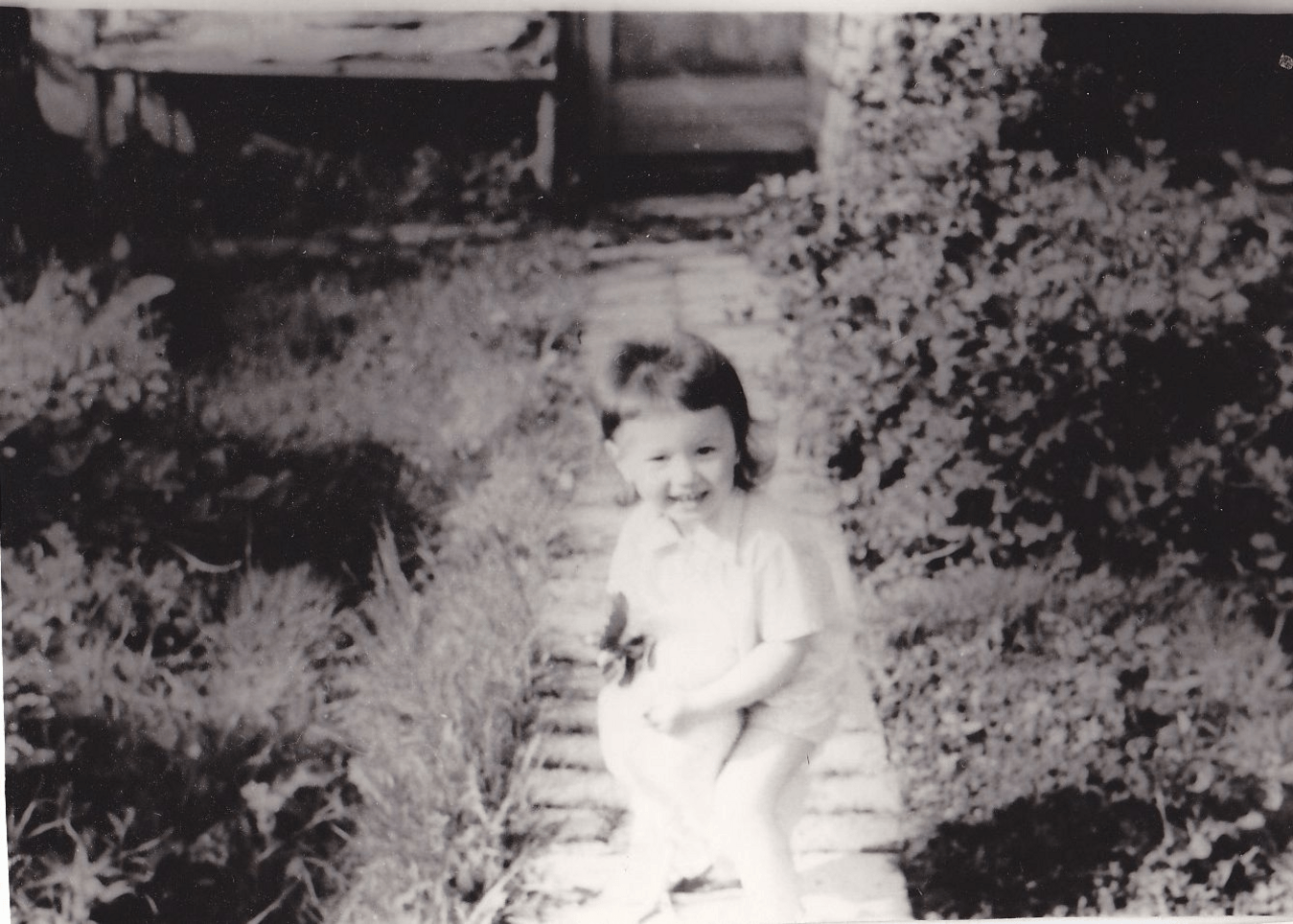 Black and white photo of a young child seated on a garden path, surrounded by greenery.