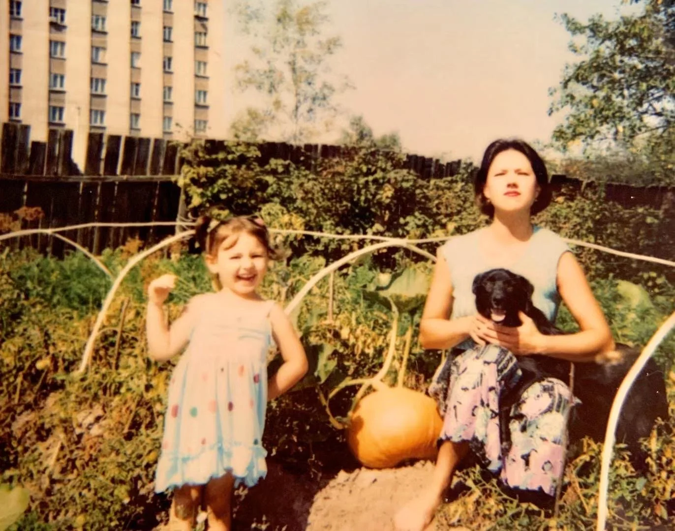 A woman holding a black puppy sits on the ground in a vegetable garden next to a large pumpkin, with a smiling young girl standing nearby. The background has a wooden fence and a tall building.