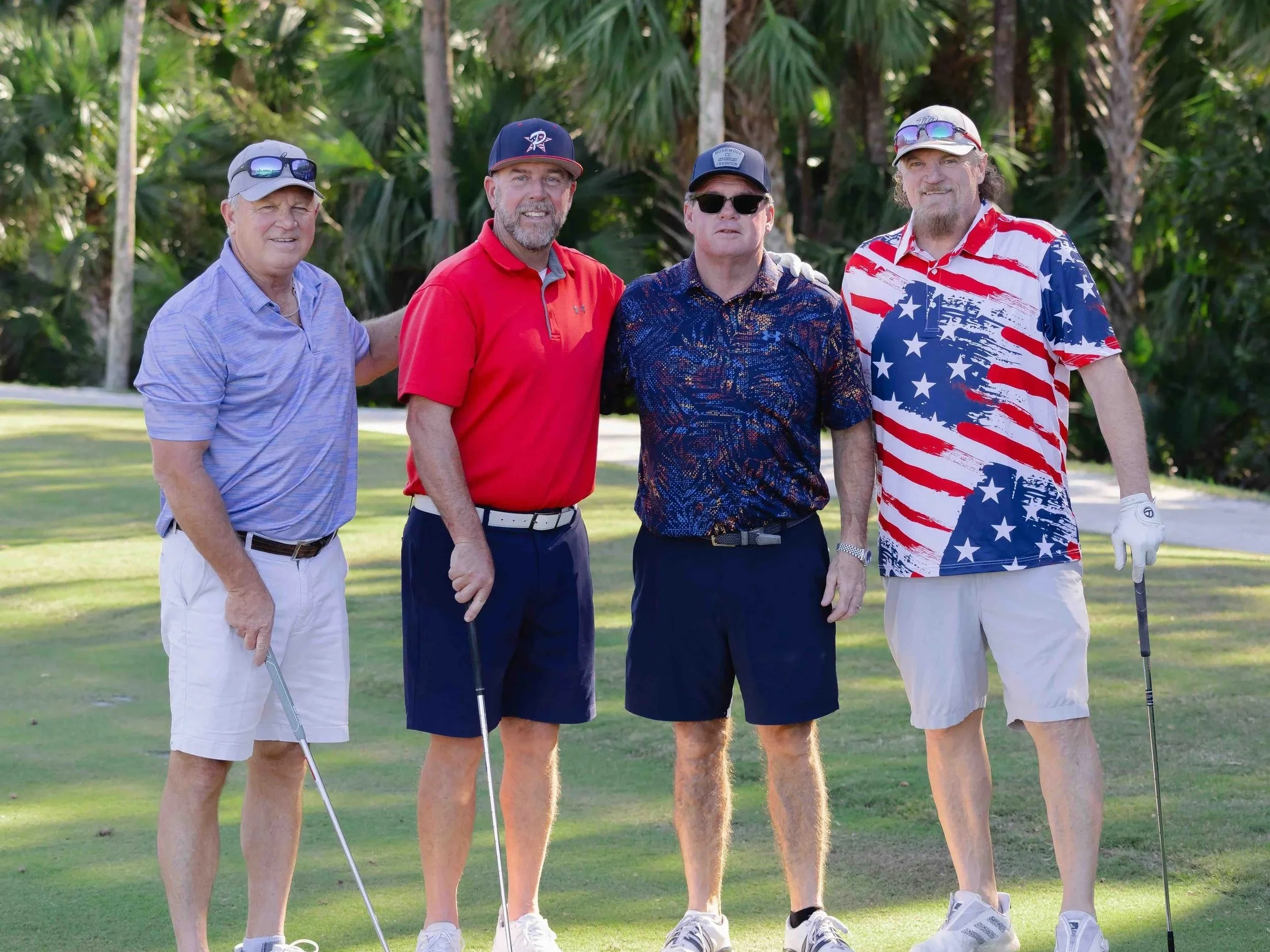 Four men standing on a golf course with trees in the background, holding golf clubs, dressed in casual golf attire.