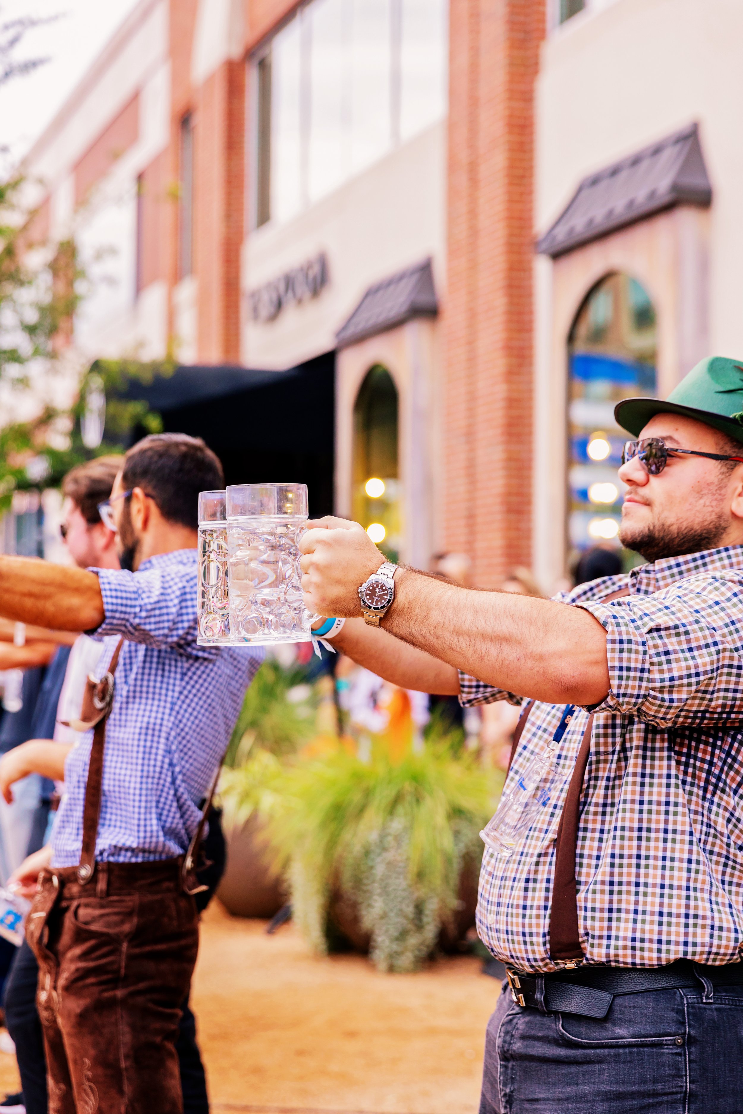 A man in a plaid shirt and sunglasses holding a large glass of beer outdoors at a festive event.