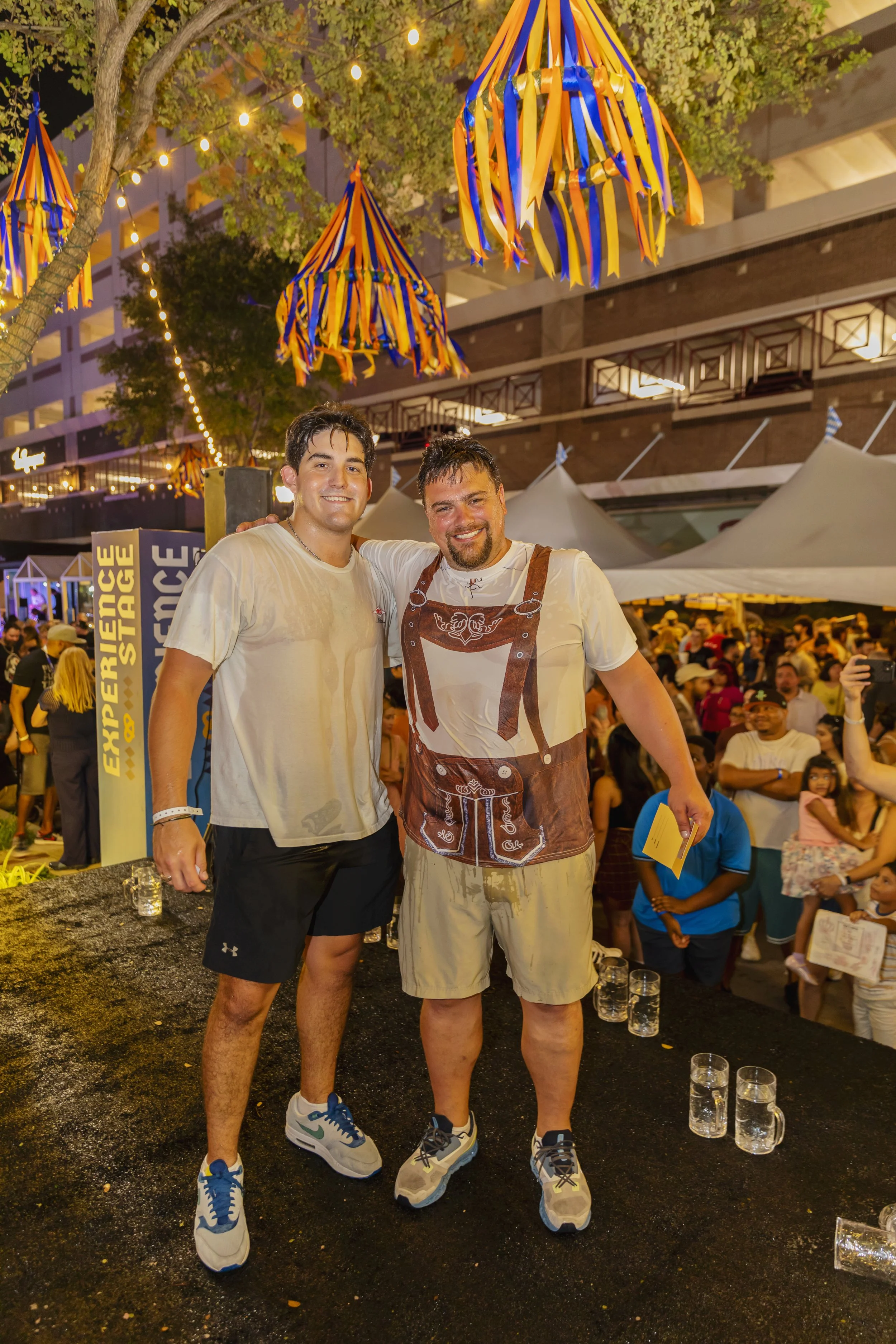 Two men smiling and posing for a photo at an outdoor event in the evening, with people and tents in the background. The setting has colorful hanging decorations and string lights.