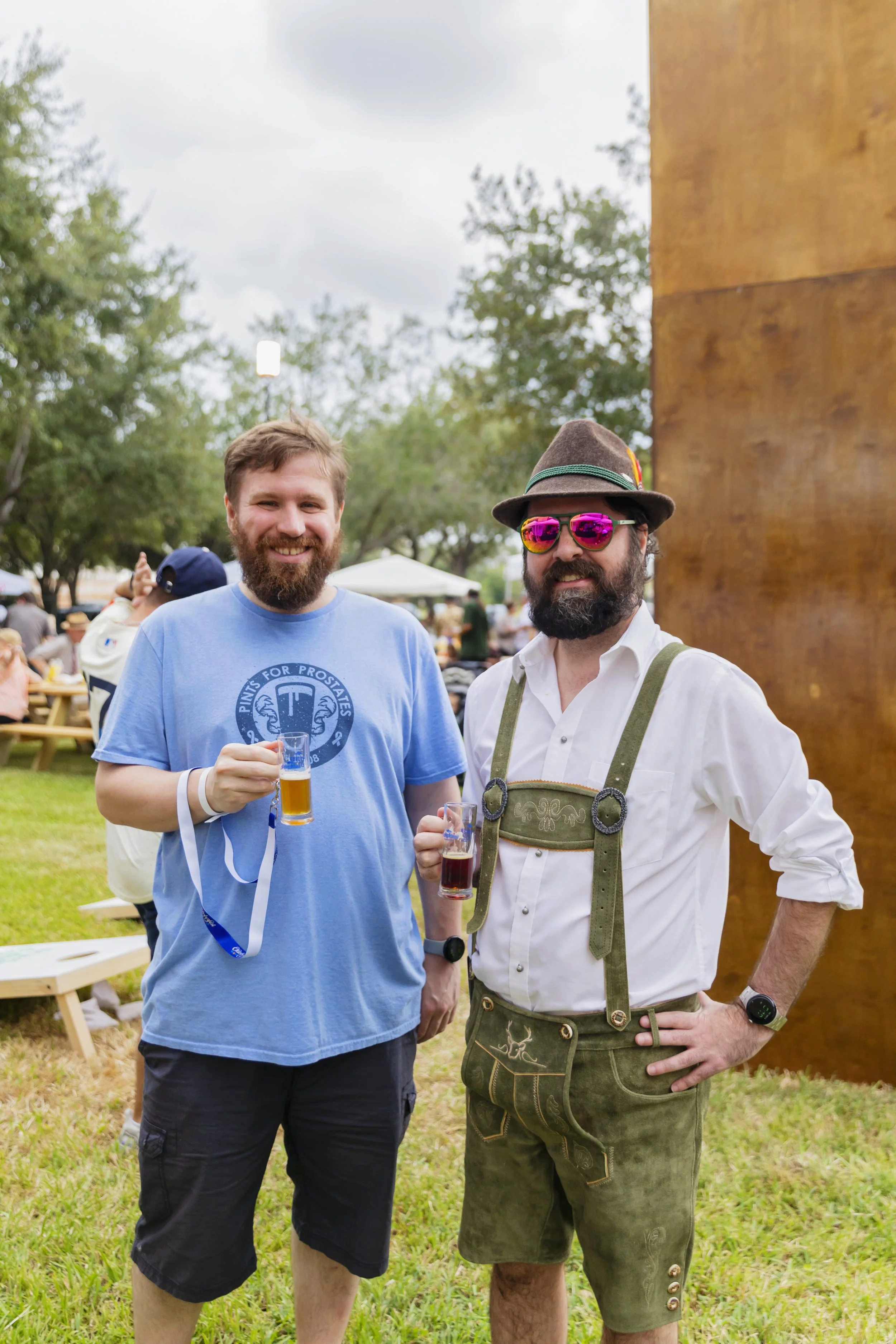 Two men standing outdoors, smiling and holding drinks at a festival or event. One wears a blue t-shirt, shorts, and a wristband, while the other is dressed in traditional Bavarian attire, including lederhosen, a white shirt, a hat, and sunglasses.