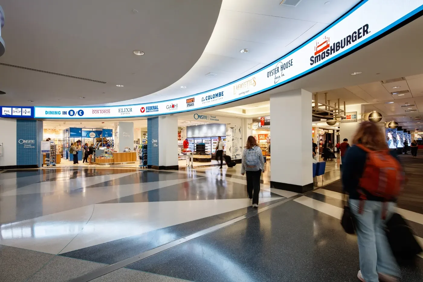 Inside an airport terminal with shops, travelers walking with luggage, and a digital directory overhead.