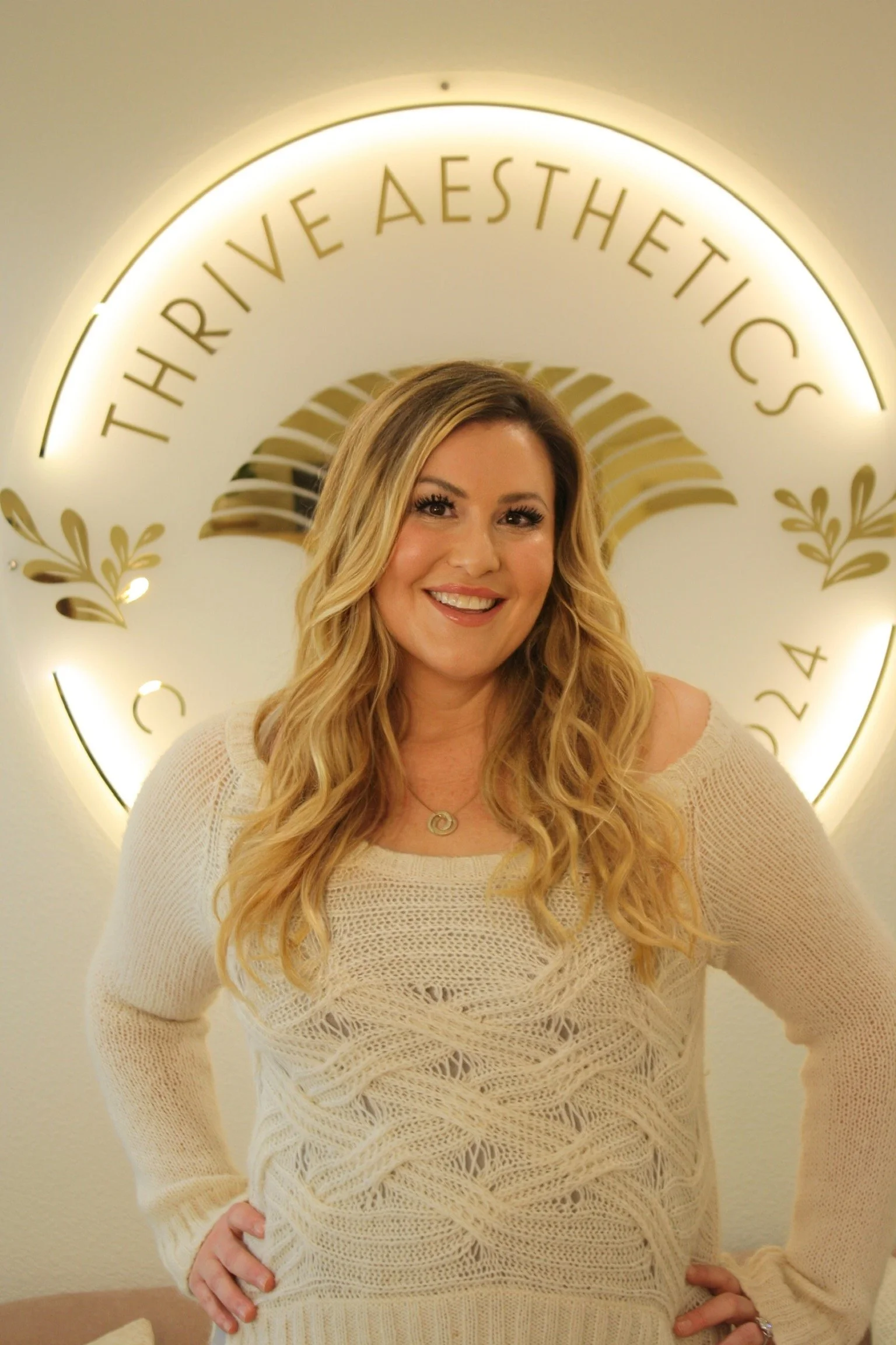A woman with long wavy blonde hair wearing a cream knit sweater, smiling, standing in front of a circular sign with the words 'THIRVE AESTHETIC' and decorative gold leaves.
