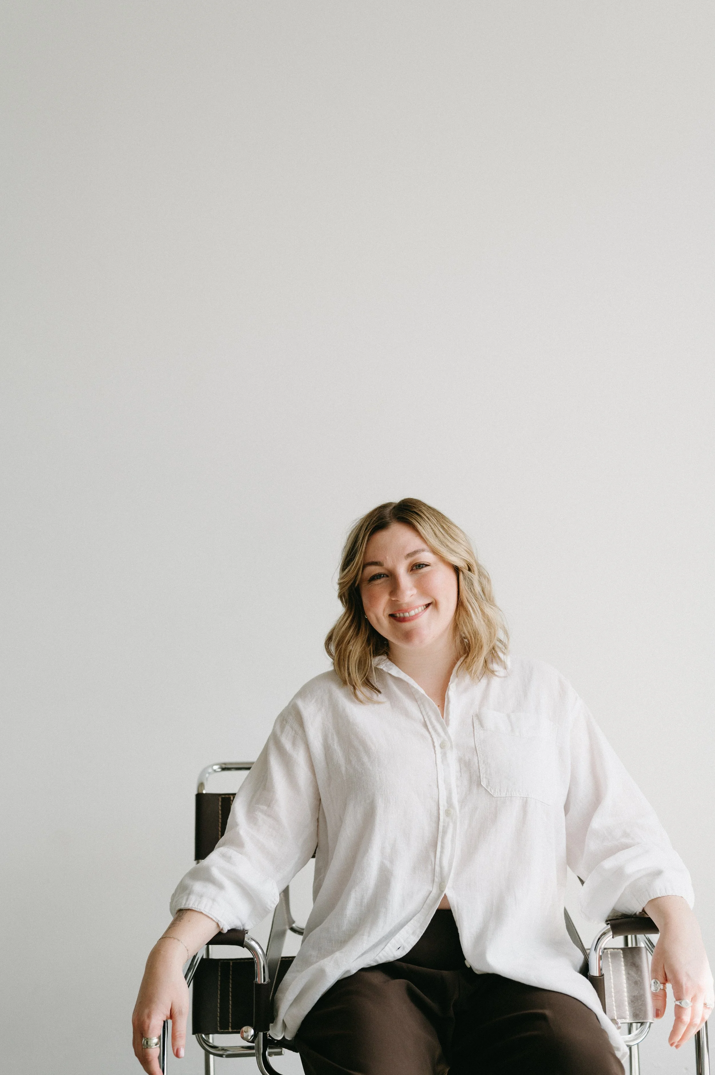 Shelby Kittinger, licensed therapist and telehealth psychologist serving Portland, Oregon and Vancouver, Washington, smiling warmly in a modern chrome chair against a light gray background