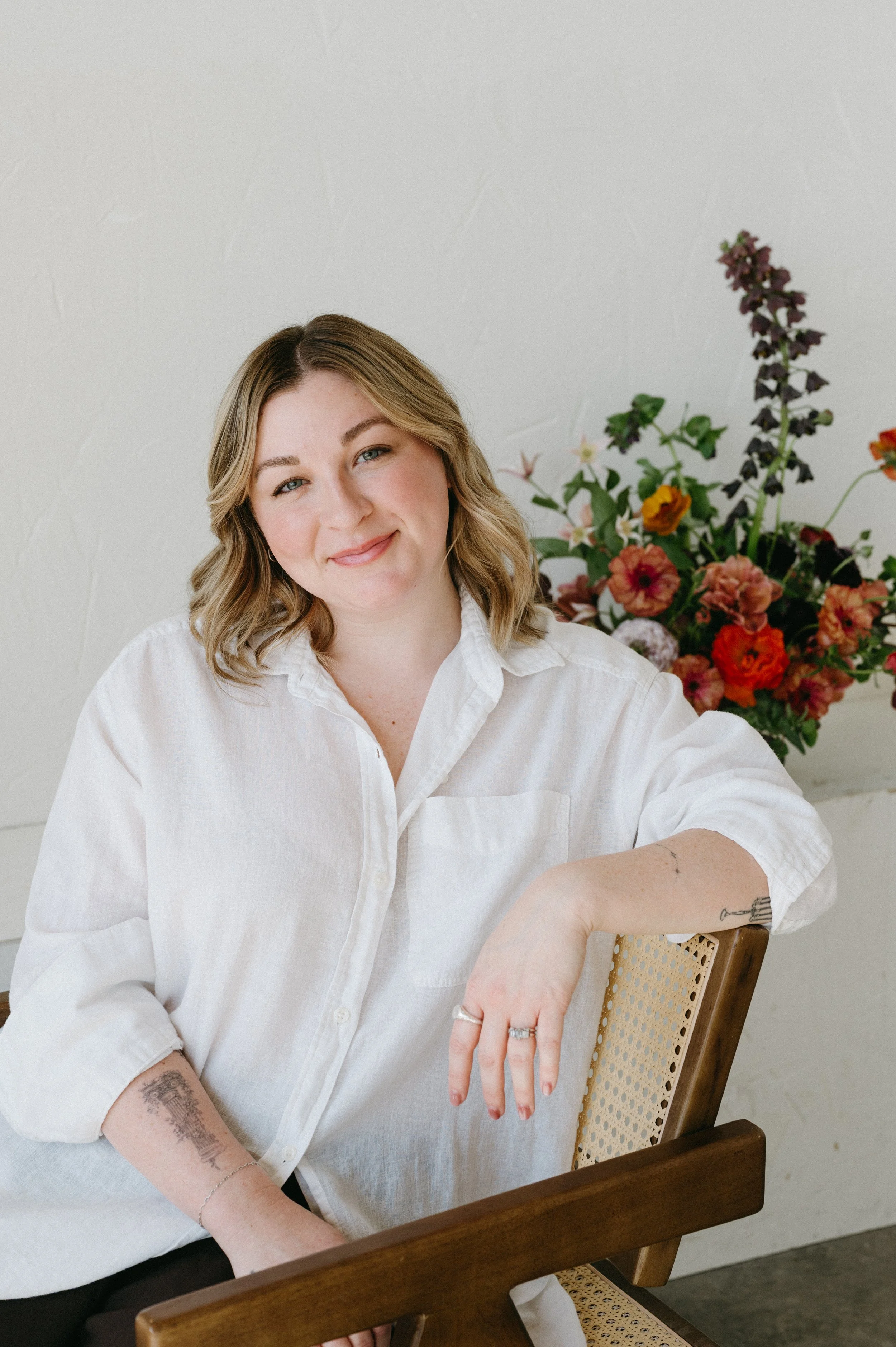 Shelby Kittinger, therapist and podcast speaker, sitting in a wooden brown chair in a white button-up shirt with a floral arrangement behind her.