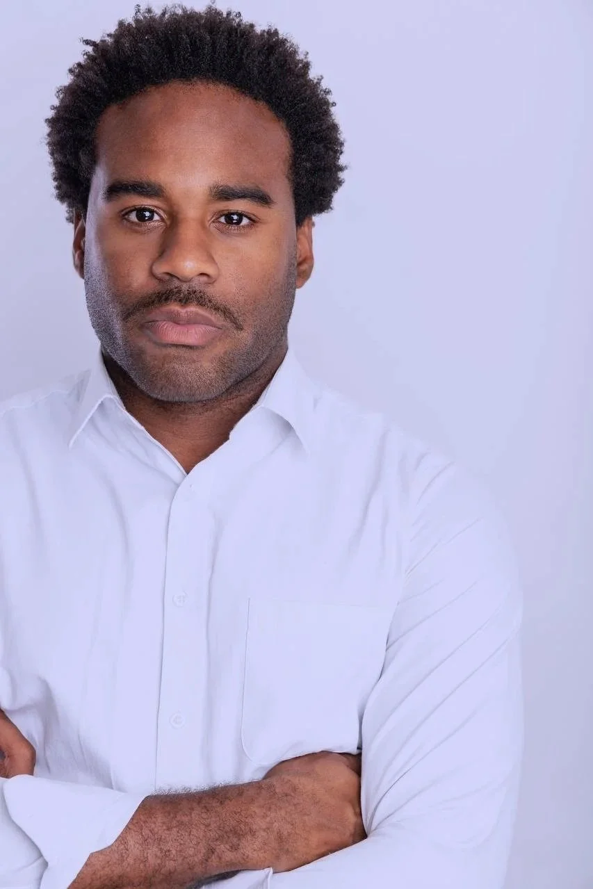 A man with dark curly hair wearing a white dress shirt, crossing his arms and looking directly at the camera against a plain light background.