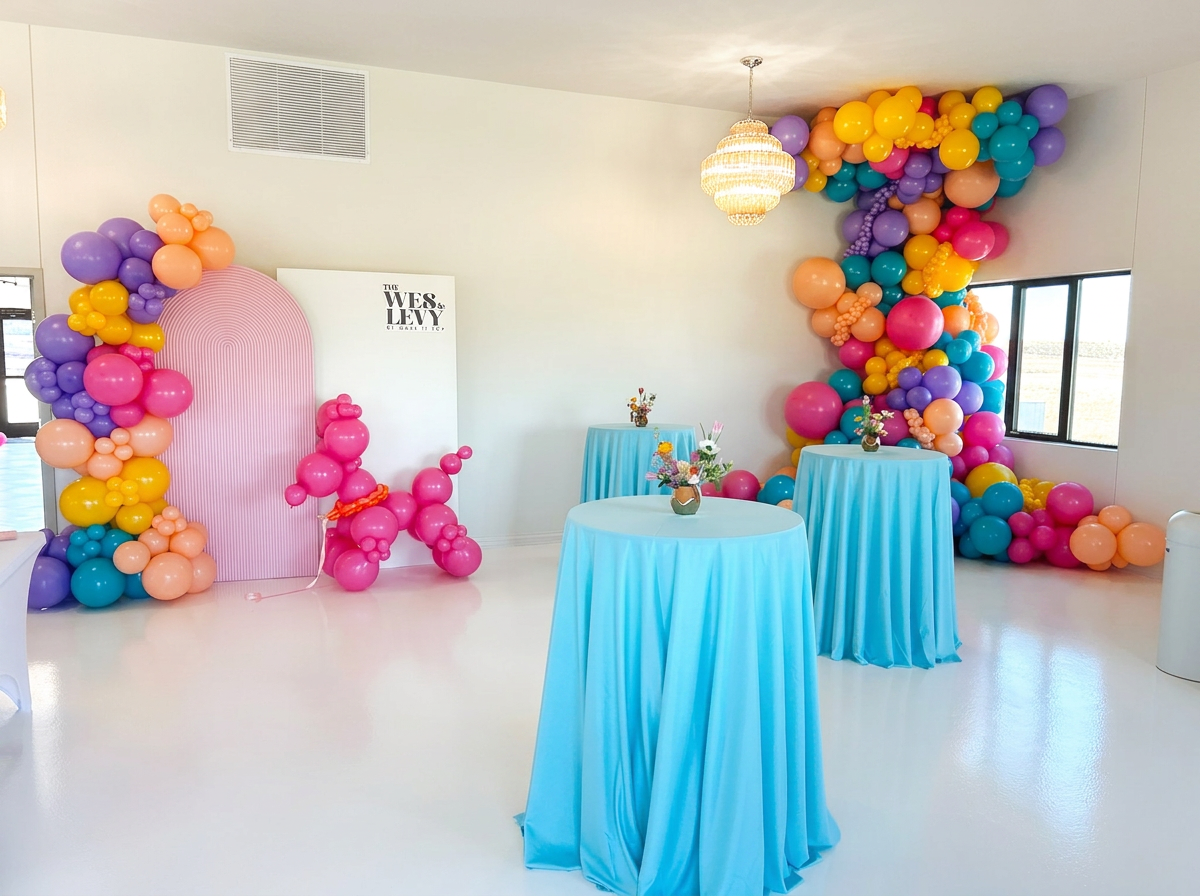 Decorative party space with balloon arch in pink, purple, yellow, blue, and orange around a pink backdrop, and a wall of balloons in similar colors near a window. Three blue-draped tables with flower centerpieces are present, and a balloon sculpture of a pink dog is near the backdrop.
