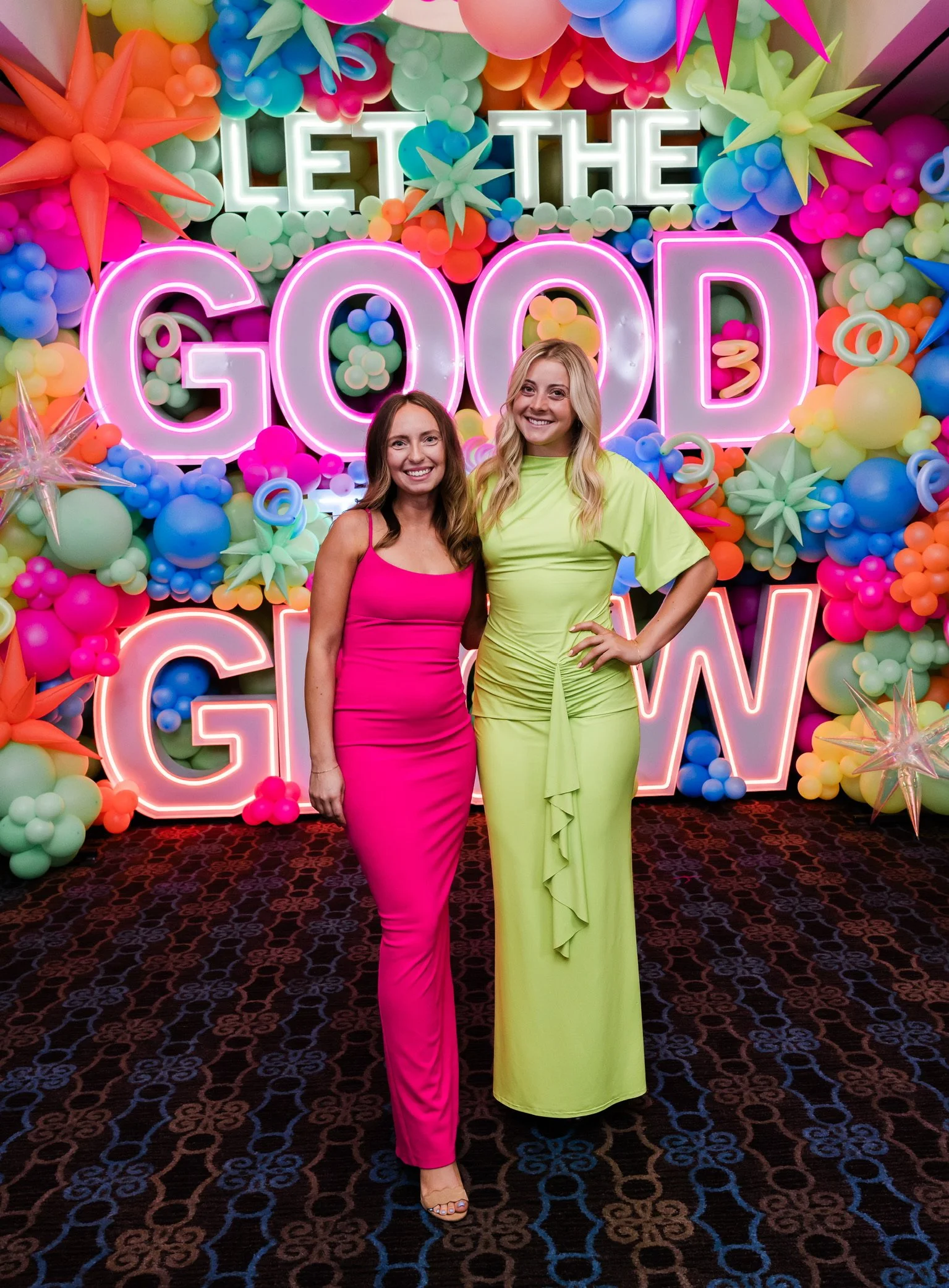 Two women smiling at the camera in front of a colorful balloon backdrop with illuminated sign saying "Let the Good times Glow".