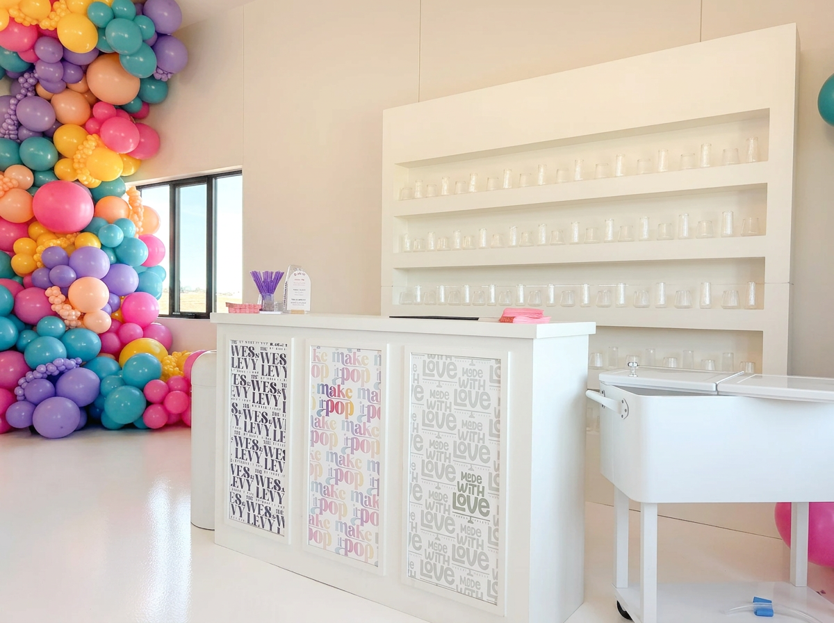 A colorful balloon decoration with pink, purple, blue, orange, and yellow balloons near a window. A white counter with three decorative panels featuring the words 'WES LEVY' and 'make pop' stands in front of a white wall with glass cups on shelves. A small white sink is positioned on the right.