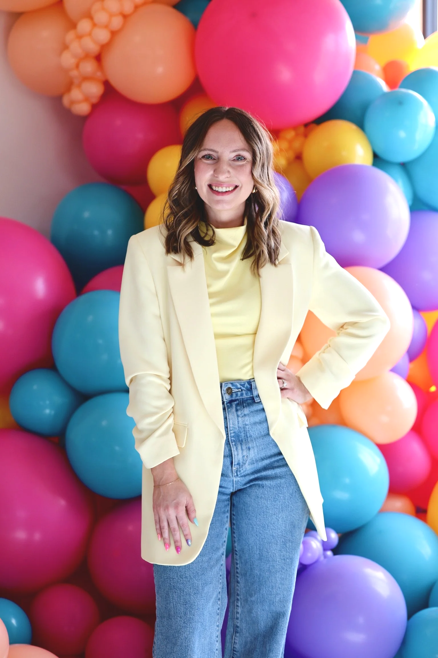 A woman with wavy brown hair wearing a light yellow blazer and blue jeans, smiling with colorful balloons in the background.