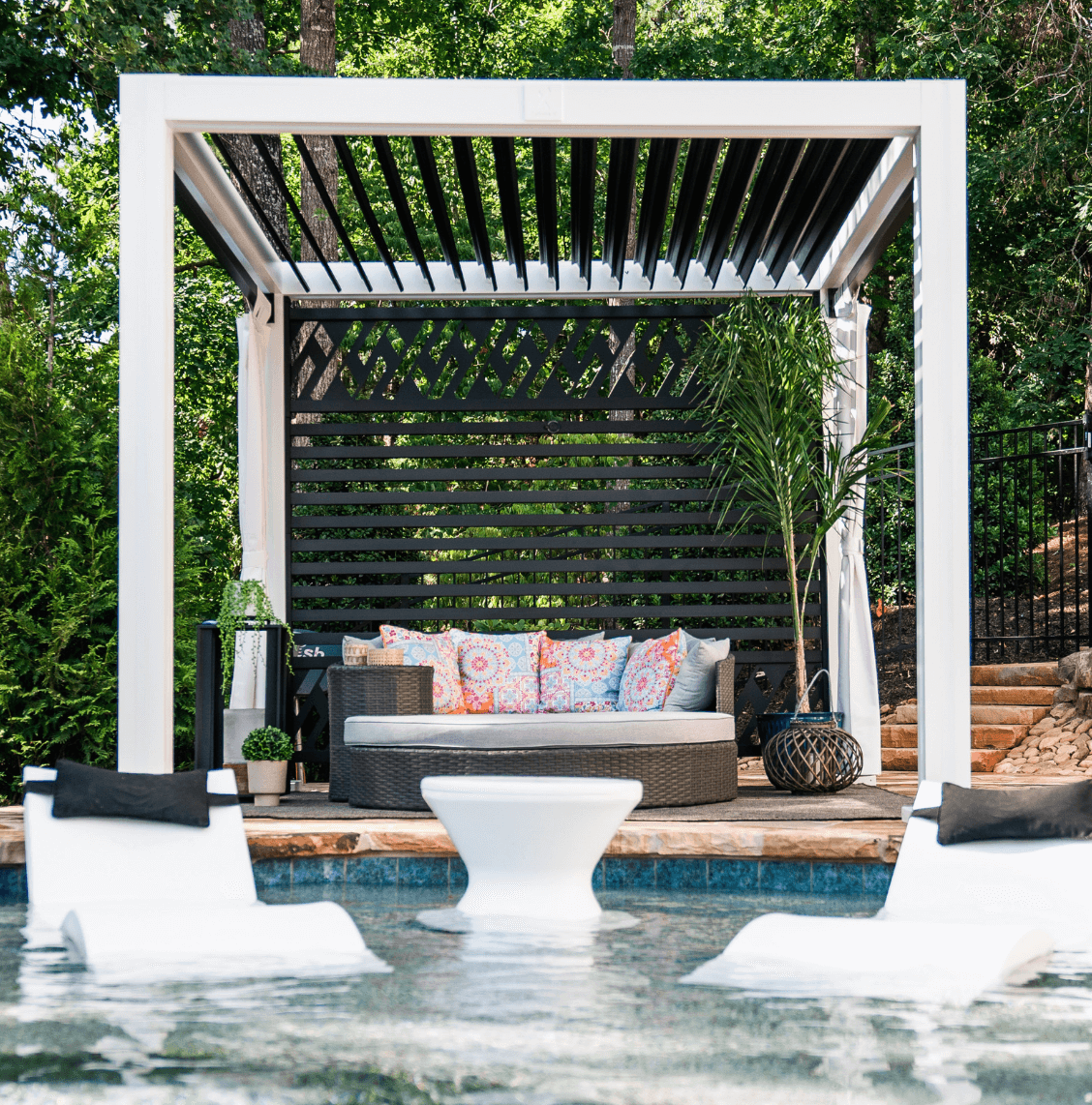 Poolside seating area with a canopy, cushioned sofa, potted plants, and pool floats in the water.