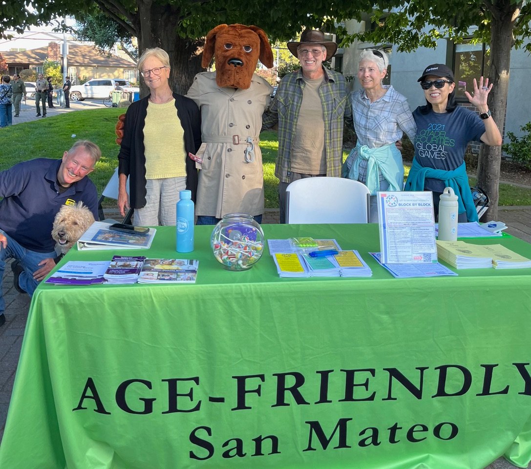 Group of six people and a dog standing behind a table covered with an 'Age-Friendly San Mateo' banner at an outdoor event. All are smiling and posing for the photo.