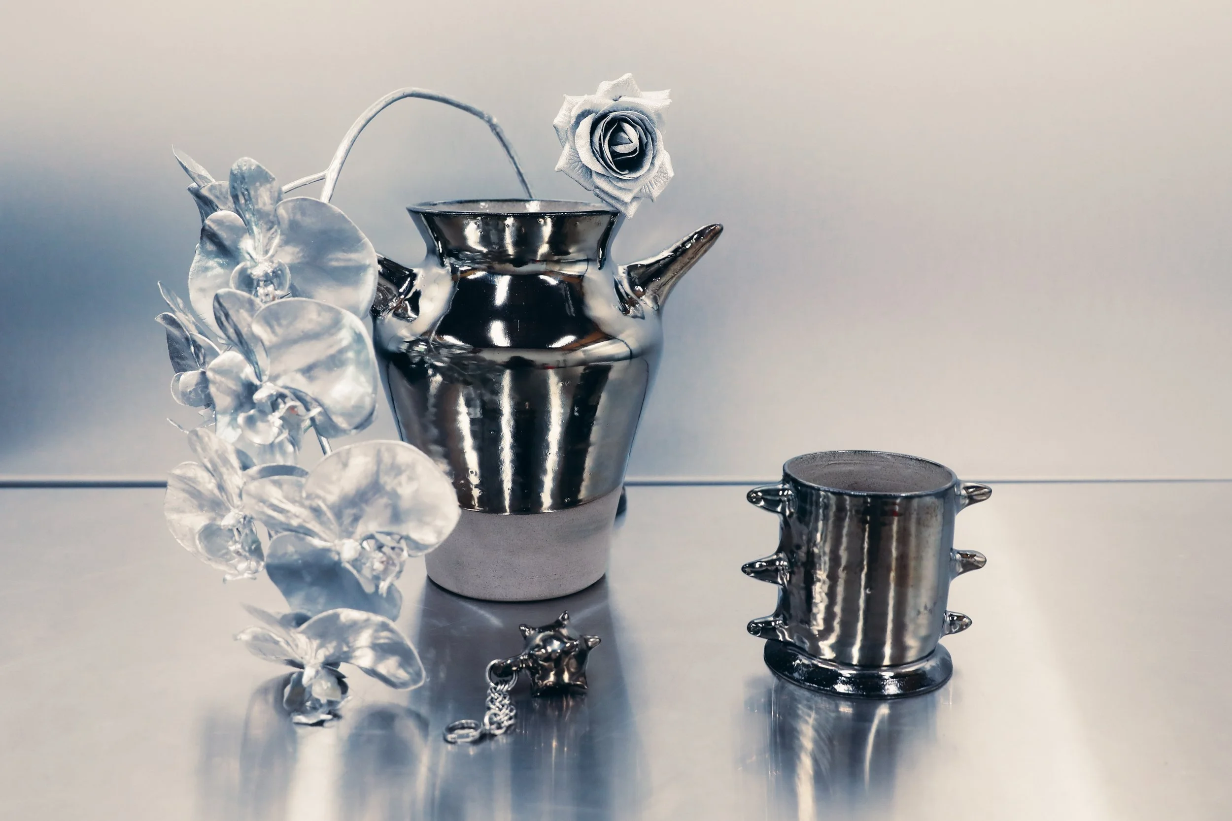 A shiny metallic teapot with a curved spout and handle, topped with a paper or fabric rose, accompanied by a small metal container and decorative metal flowers, all reflected on a smooth surface.
