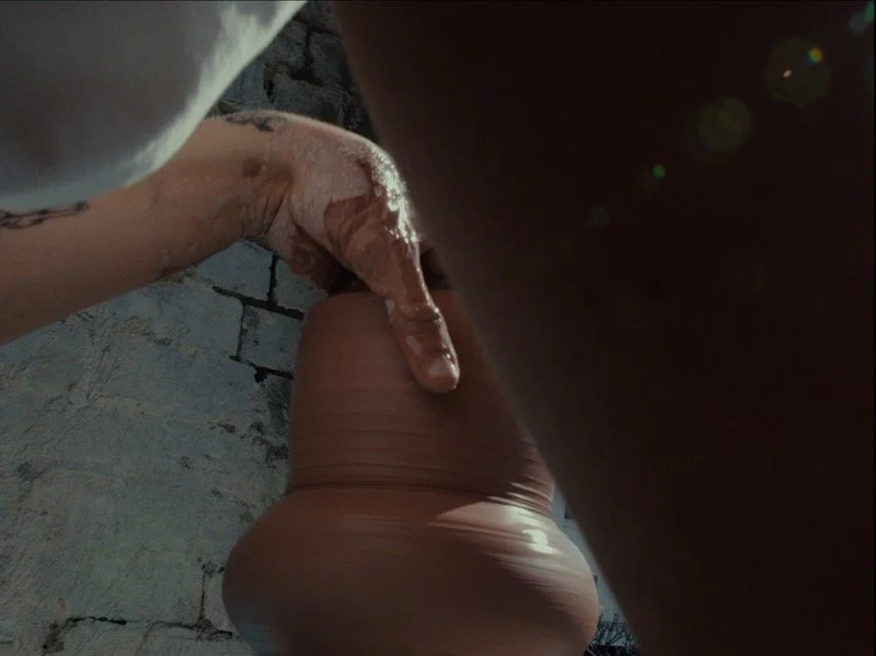 A person is shaping a clay vase on a pottery wheel with their hand. The background shows a brick wall and a bright light of sunlight.
