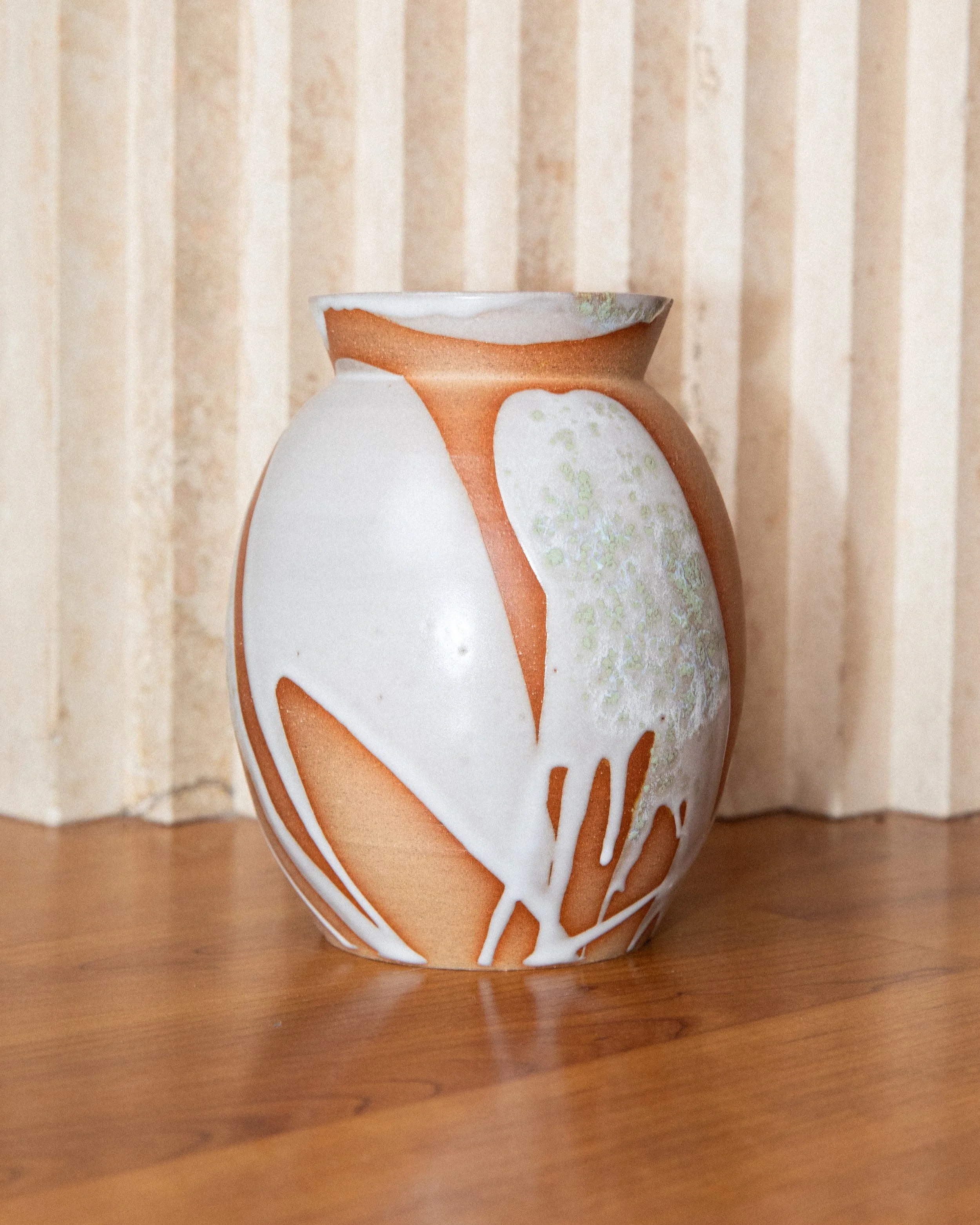 Ceramic vase with a white and orange abstract pattern, placed on a wooden surface with a beige striped wall in the background.