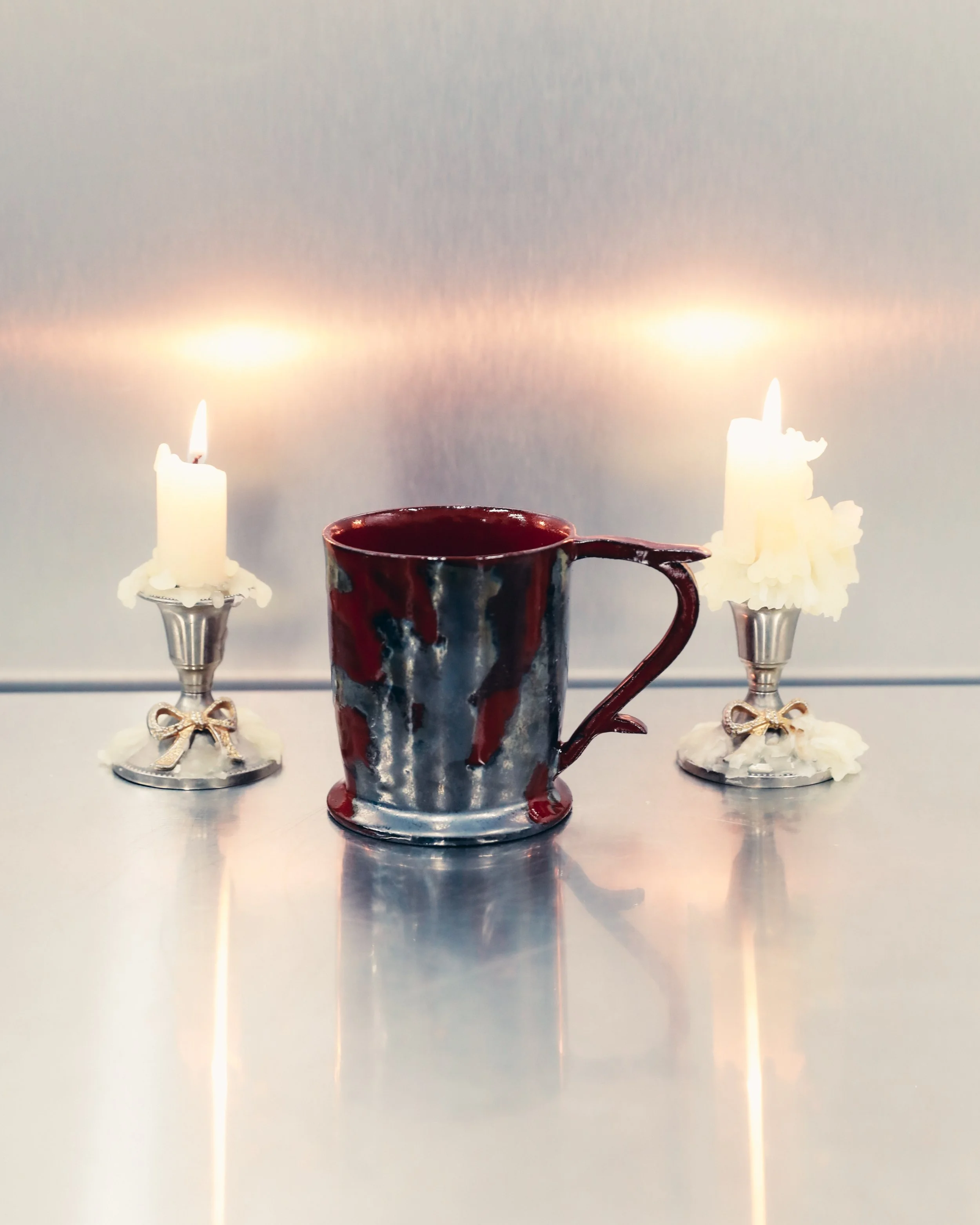 A red and black striped ceramic mug with a decorative heart-shaped handle, flanked by two white candles with melted wax and metal candle holders with bows, all placed on a reflective surface against a gray background.