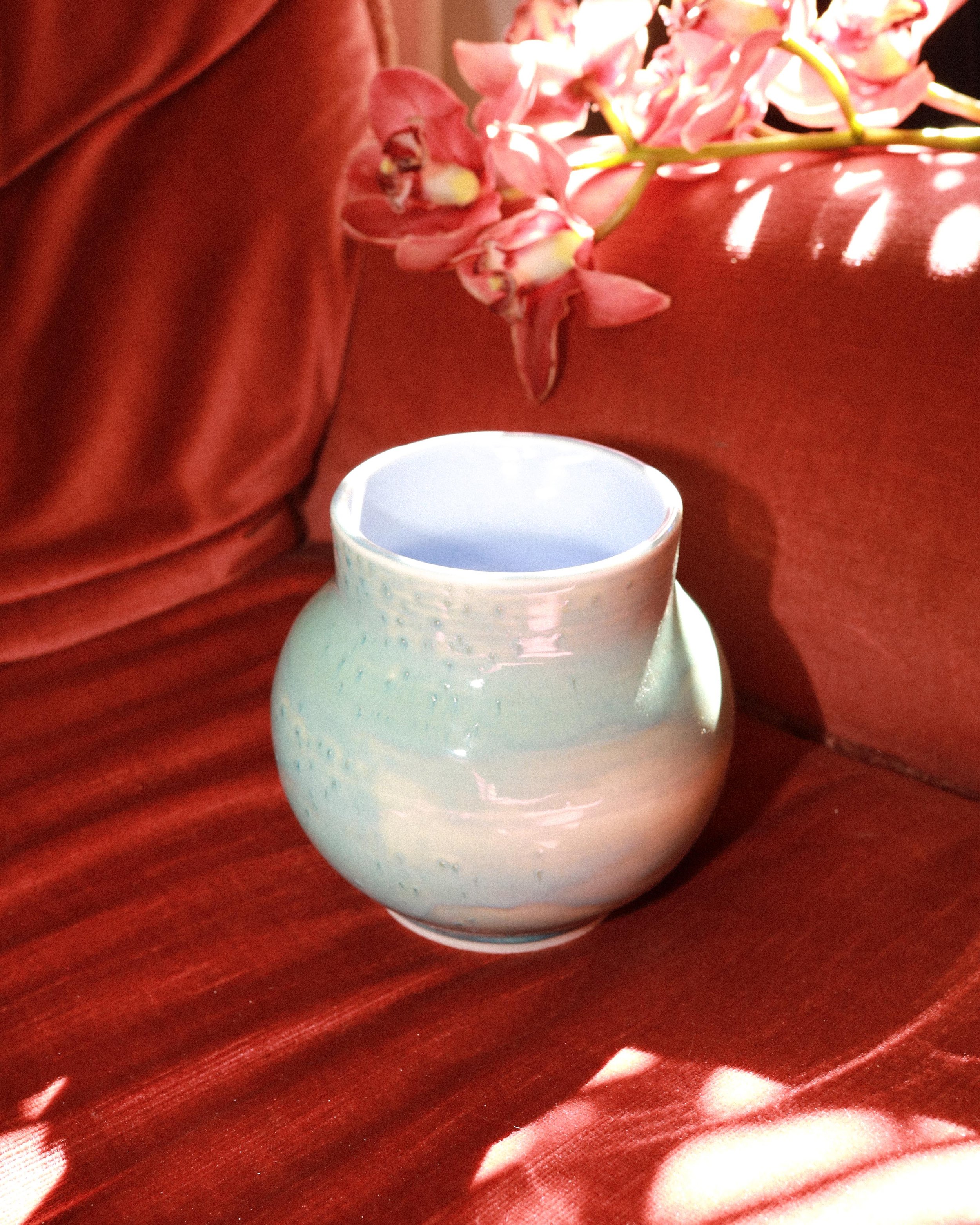 A white ceramic mug on a red satin cloth. Pink orchid flowers rest on the cloth next to the mug. Sunlight creates reflections on the satin fabric.
