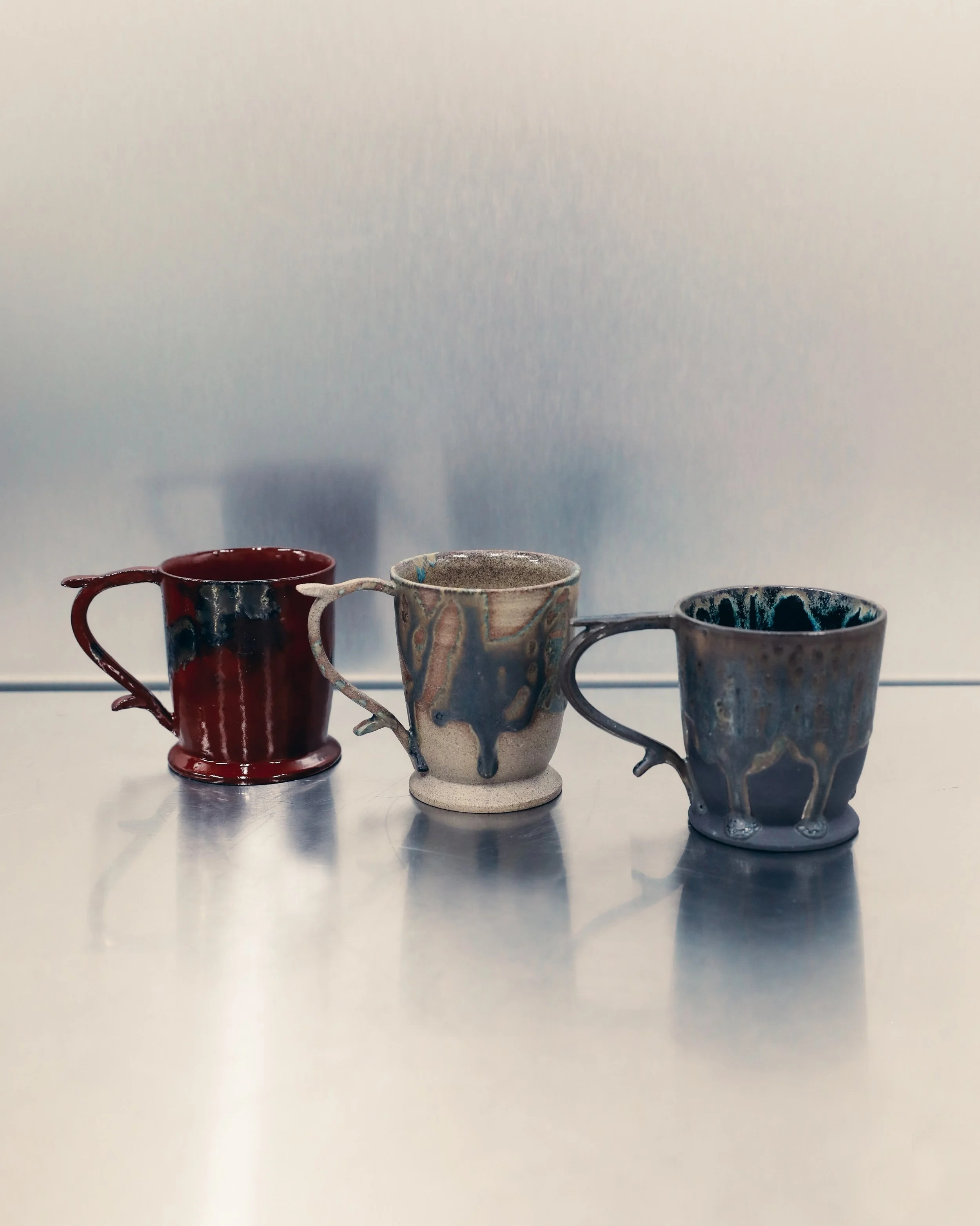 Three ceramic mugs with different colors and patterns are lined up on a reflective surface against a plain white background.