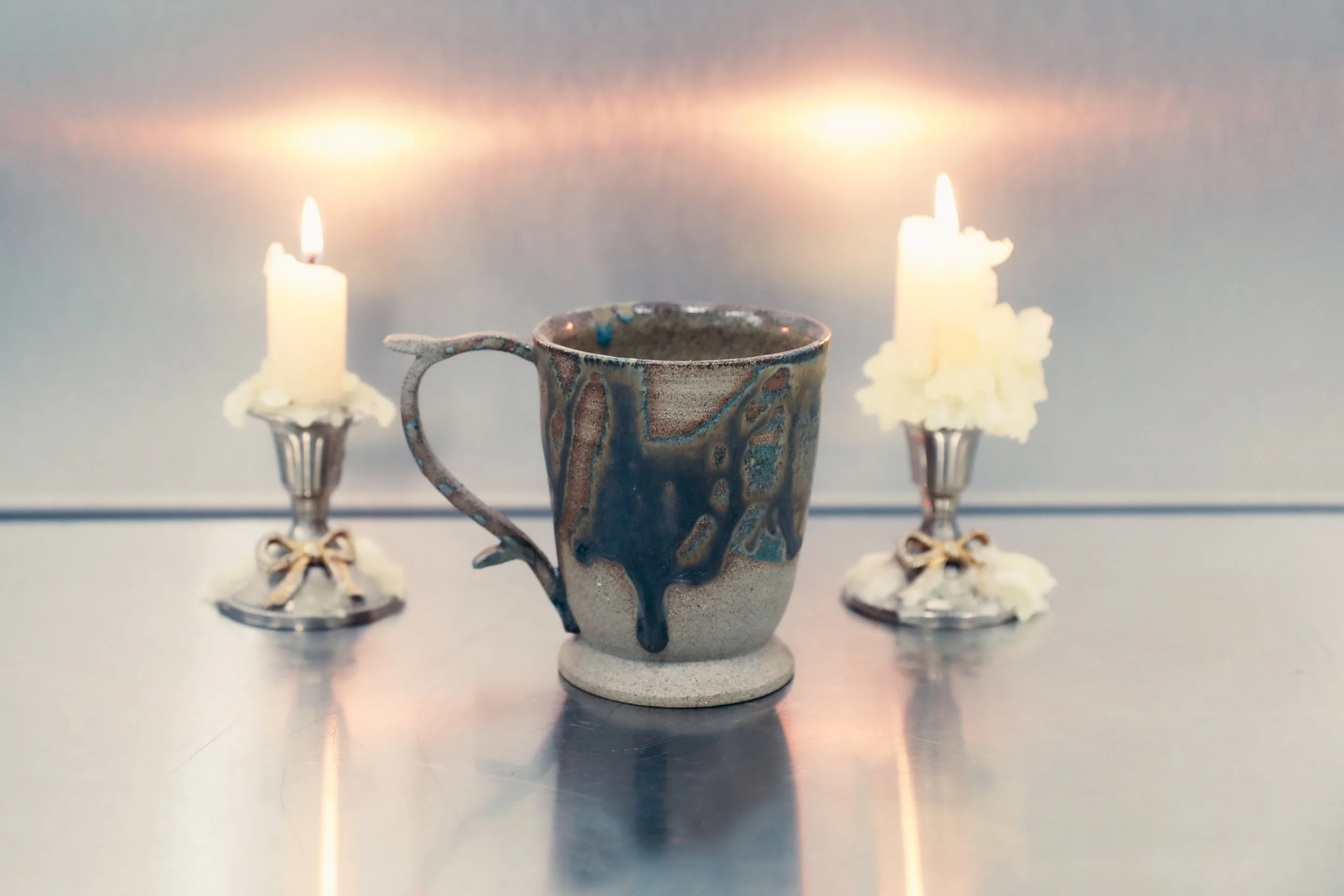A ceramic cup with blue and brown glaze in front of two lit candles with melted wax, all on a reflective metallic surface.