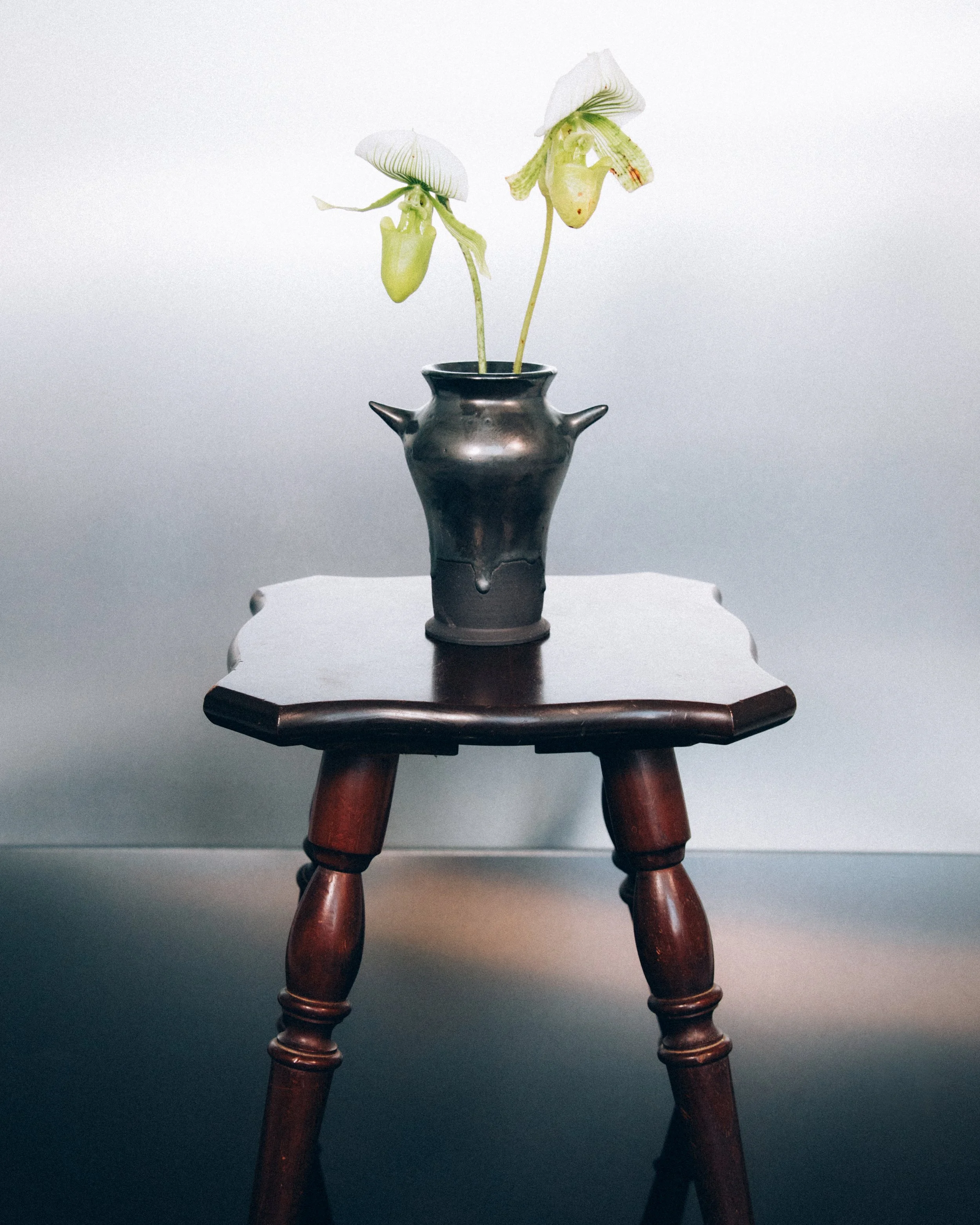 A black ceramic vase with two orchid stems with white flowers, placed on a small dark wooden table against a plain light gray background.