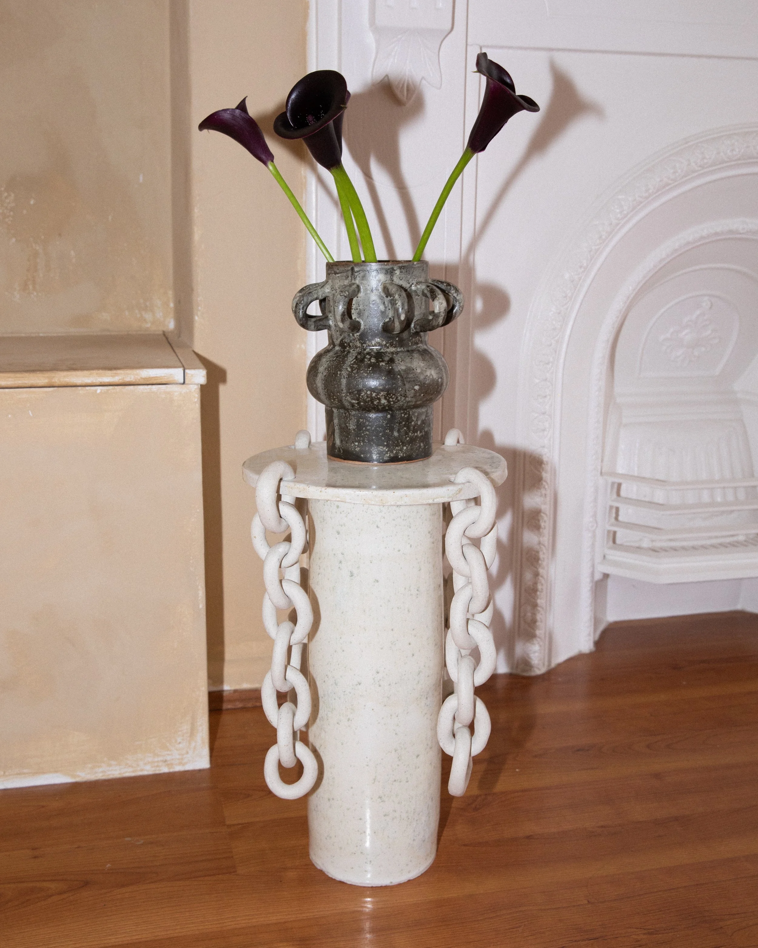 A white ceramic vase on a stand with three dark purple calla lilies. The stand has decorative white chains hanging from it. The background includes a beige wall, a part of a beige furniture piece and a white decorative wall panel or fireplace mantle.