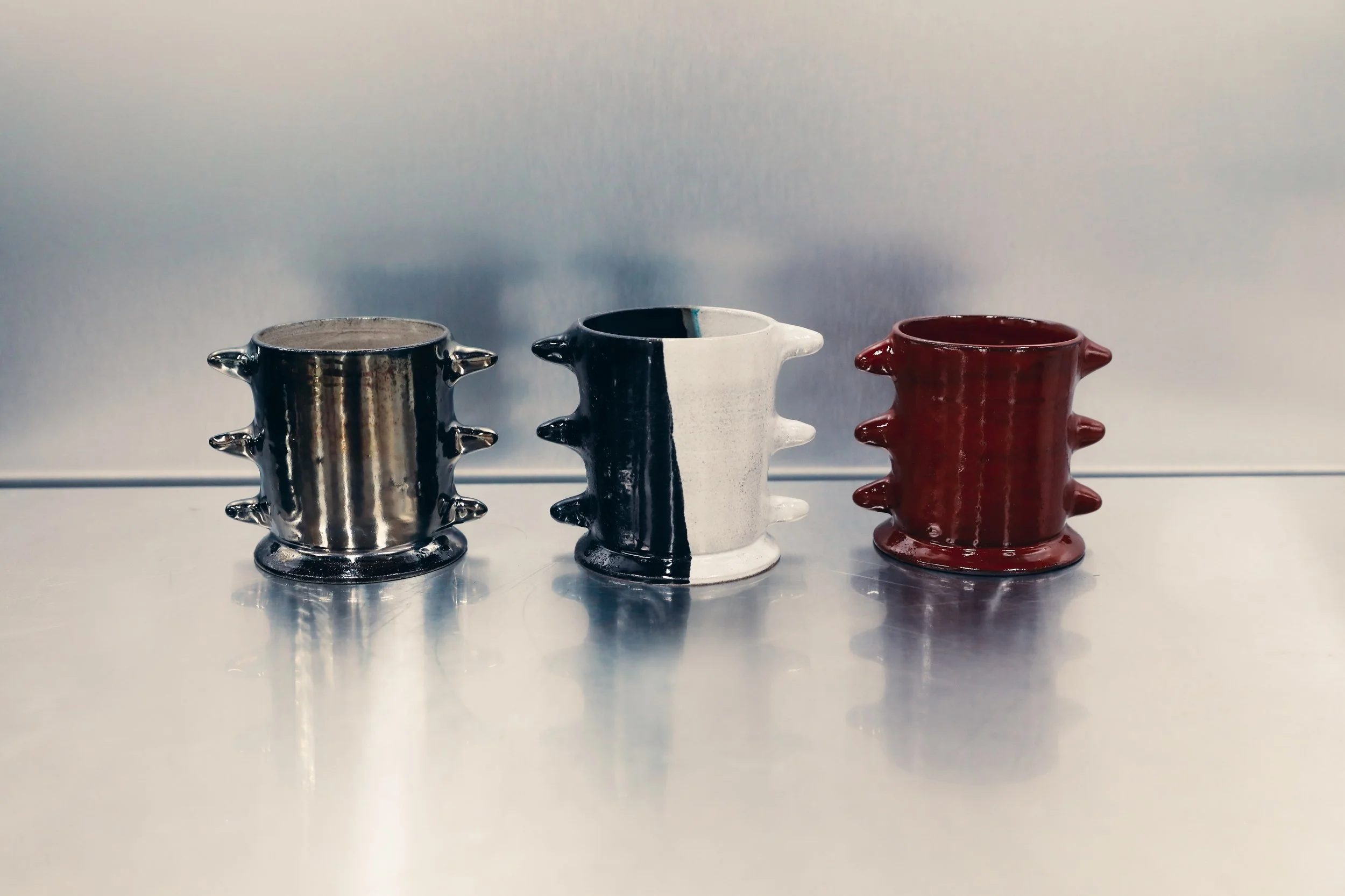 Three small ceramic cups with spike-like protrusions, each painted in different colors: one metallic silver, one black and white, and one red, arranged in a row on a reflective surface against a plain background.