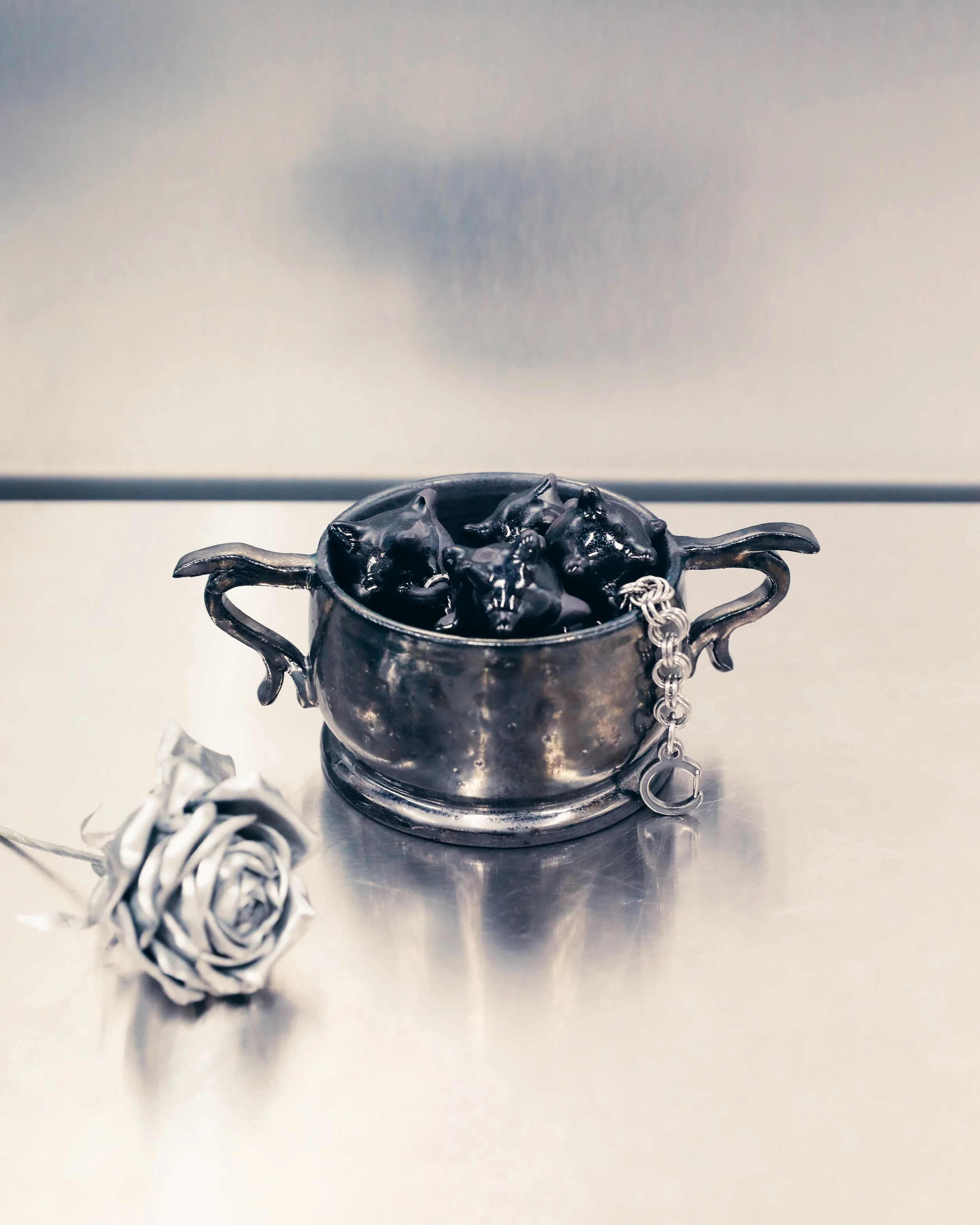 A black metal bowl with ornate handles, the bowl appears to contain an object that resembles a black sculpture of a horse or a similar animal. A silver chain is attached to the bowl, draping over its side. The background is plain and gray.