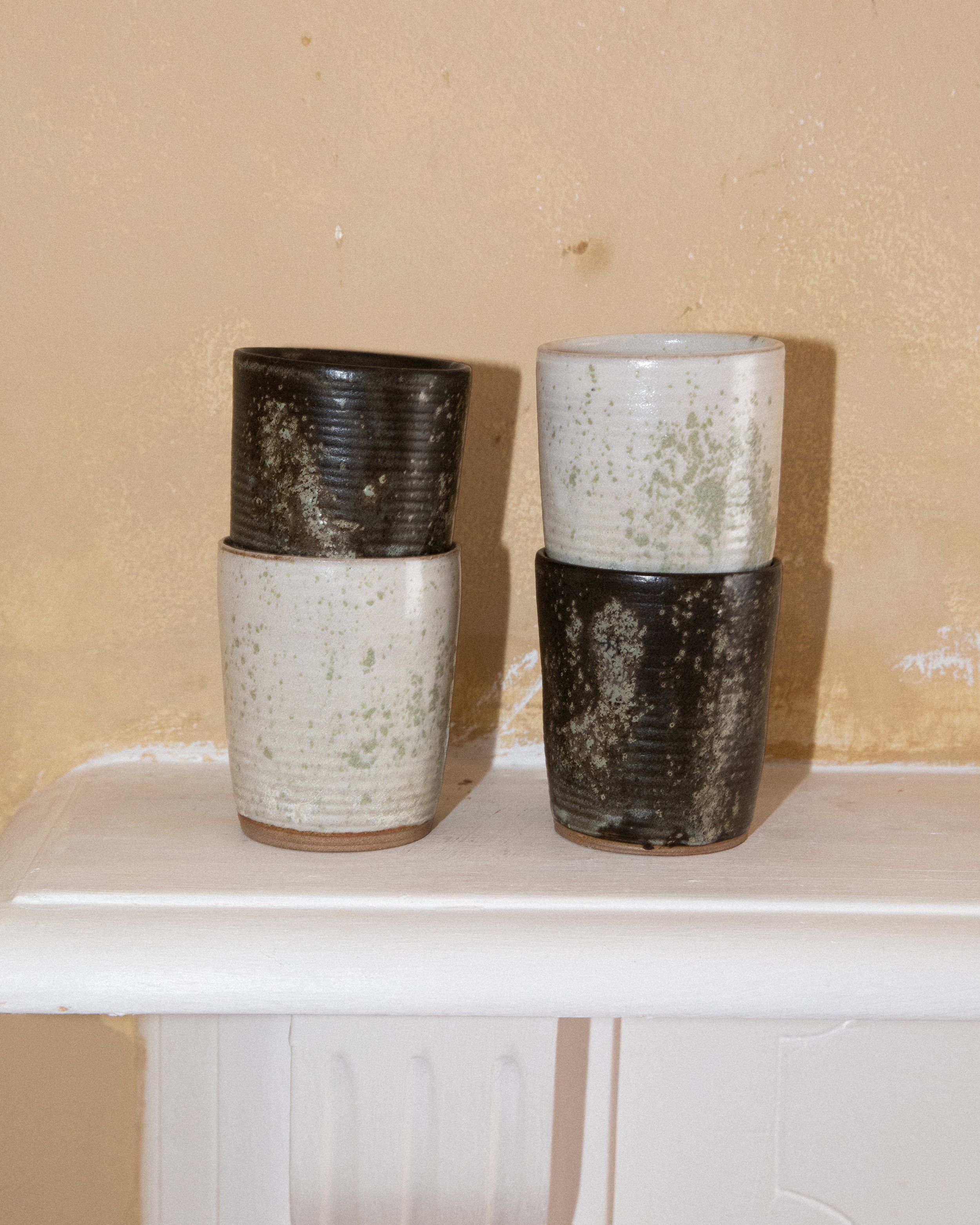 Six ceramic cups, three black and three white with green speckles, stacked on a white shelf against a beige wall.