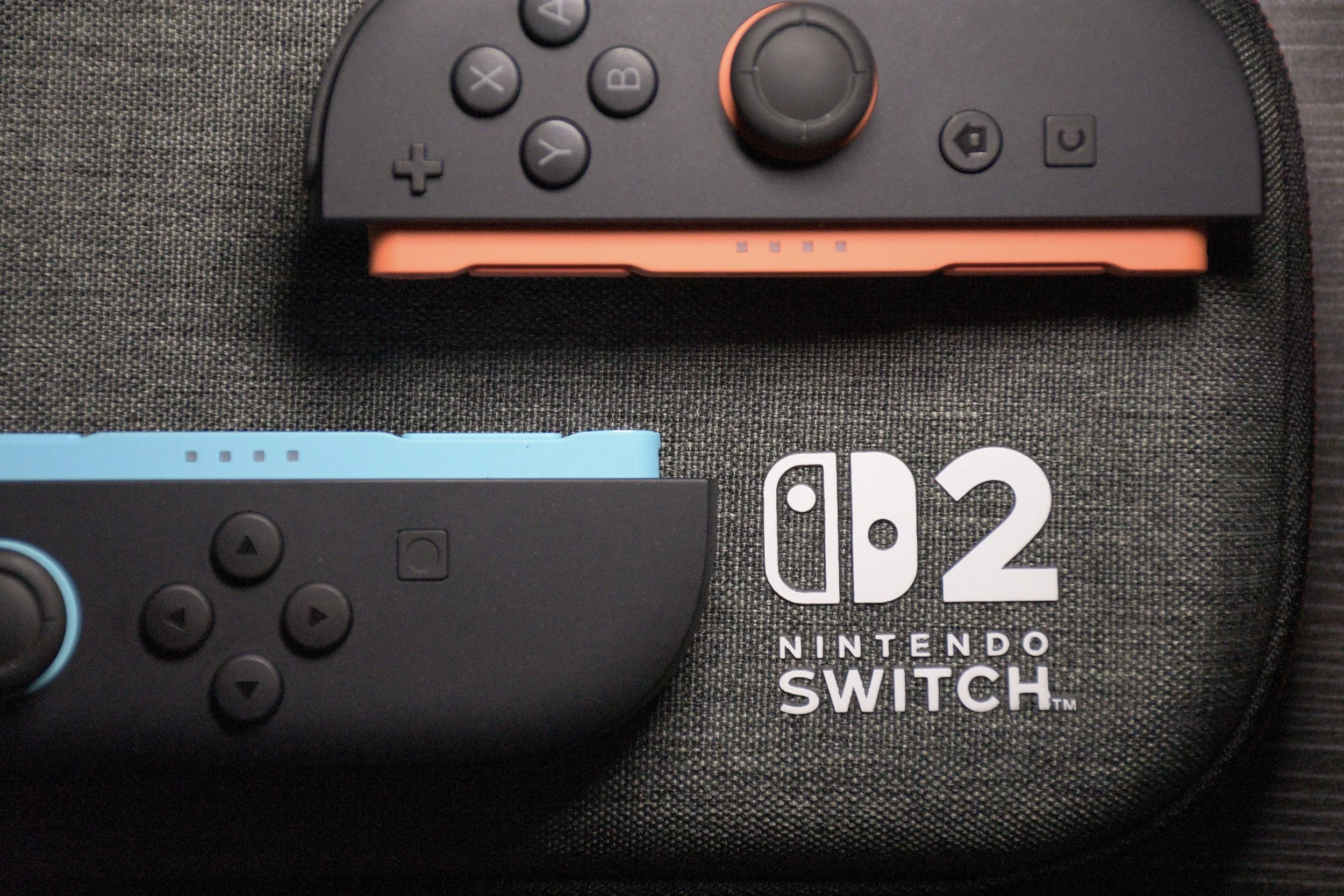 A Nintendo Switch gaming console case with a black fabric texture, featuring a white Nintendo Switch logo, the number 2, and the word 'Nintendo Switch' printed below. Two Nintendo Switch controllers, one with a pink and black color scheme and the other with a black and blue color scheme, are placed on top of the case.