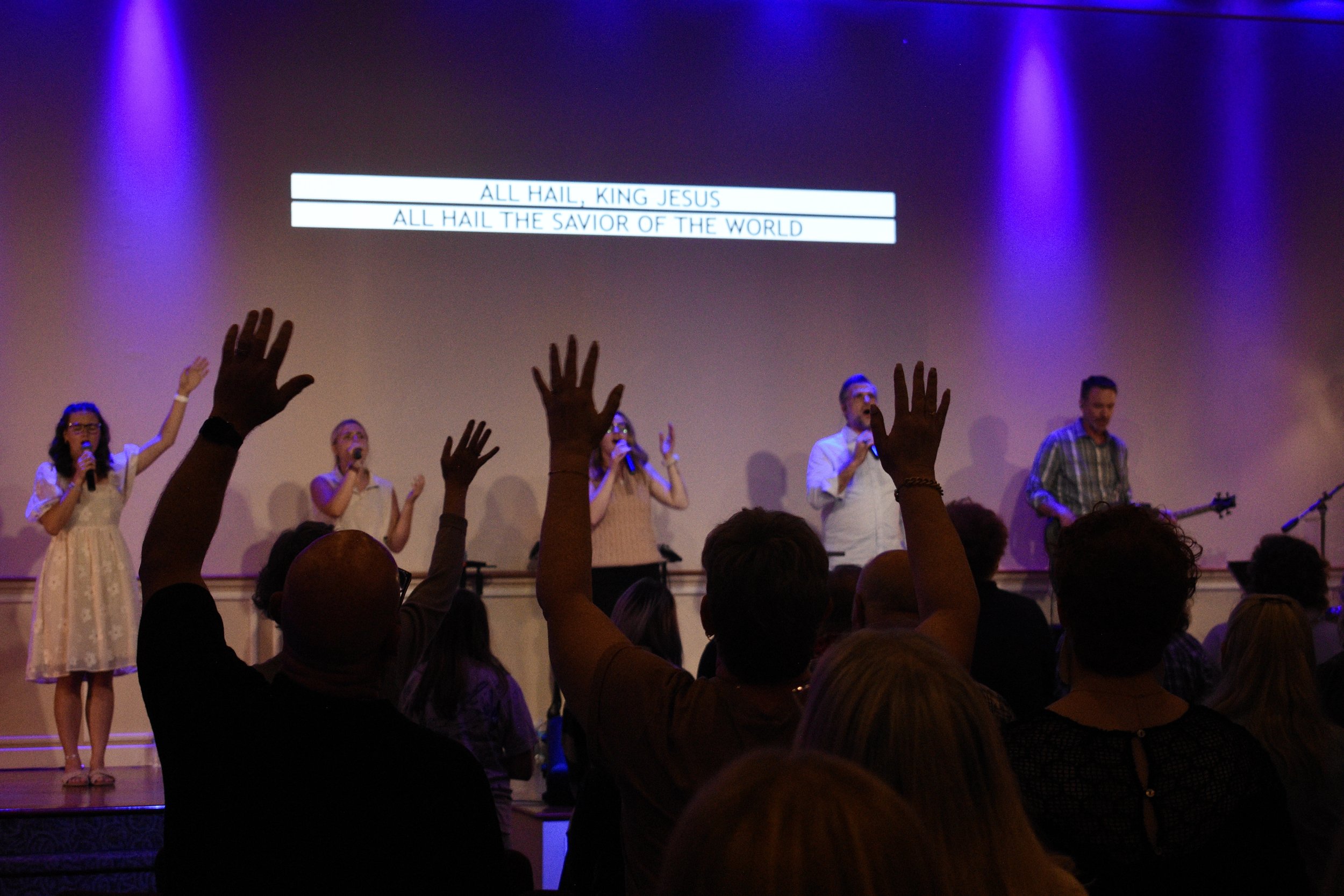 Church congregation singing with raised hands during worship, with lyrics on screen reading 'All hail, King Jesus' and 'All hail the savior of the world.'