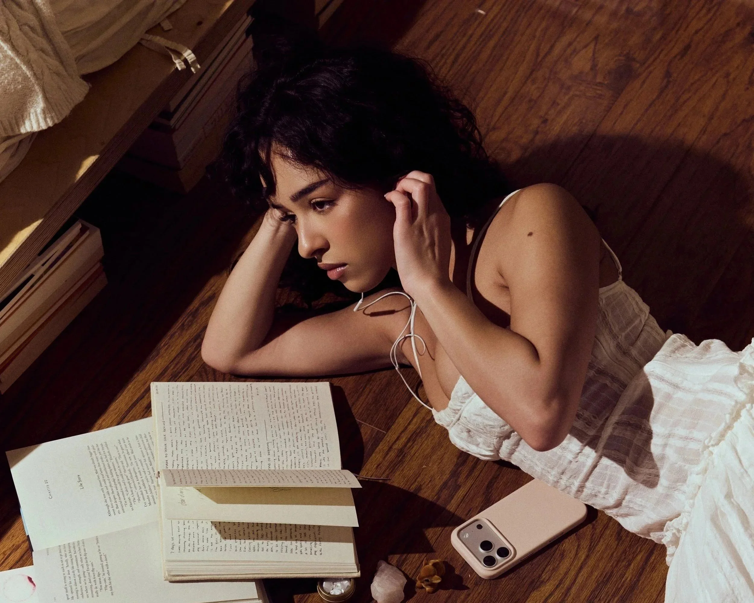 A woman lying on her side on a wooden floor, reading an open book with multiple sticky notes. She has dark, curly hair, is wearing a light-colored sleeveless top and a ruffled skirt, and has earphones in her ears. There is a smartphone and some small objects on the floor next to her.