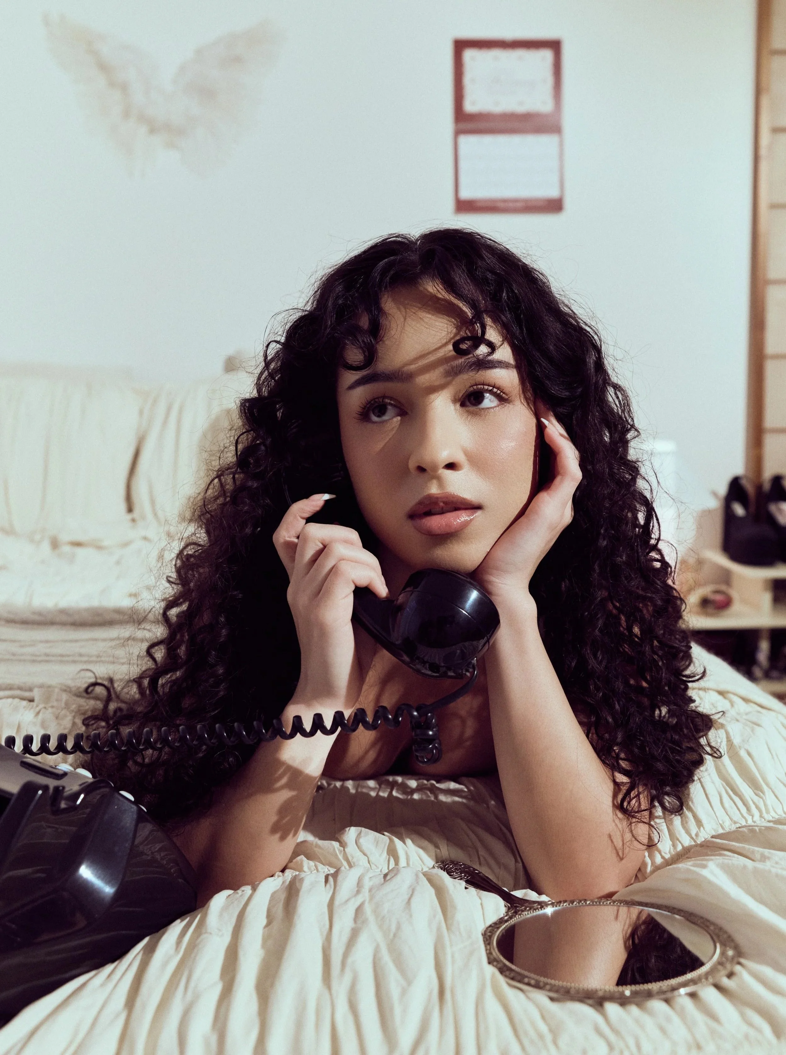 A woman with curly hair lying on a bed, holding a black telephone receiver to her ear, looking thoughtfully to the side with a mirror in front of her.