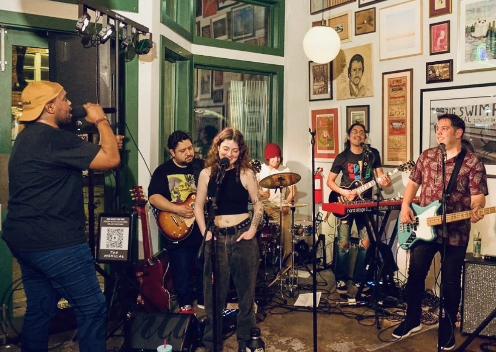 A band performing in a cozy, art-filled venue with five musicians, including a singer, guitarist, keyboardist, bassist, and drummer, with various framed artwork on the wall behind them.