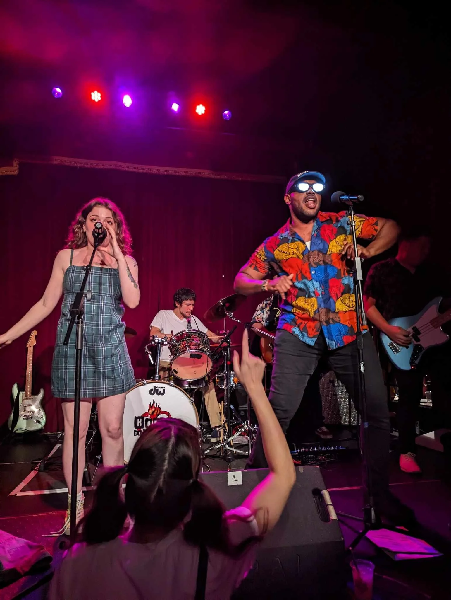 A band performing on stage with a female singer, a male rapper, a drummer, and a guitarist in front of a red curtain, colorful stage lights overhead, and an audience member raising a hand.