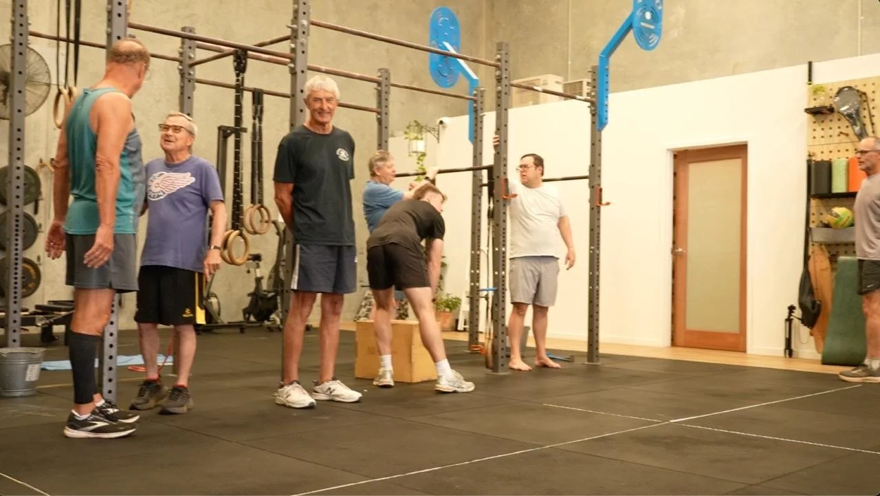 Group of six older adults standing in a gym smiling, some talking, in workout clothes, near fitness equipment