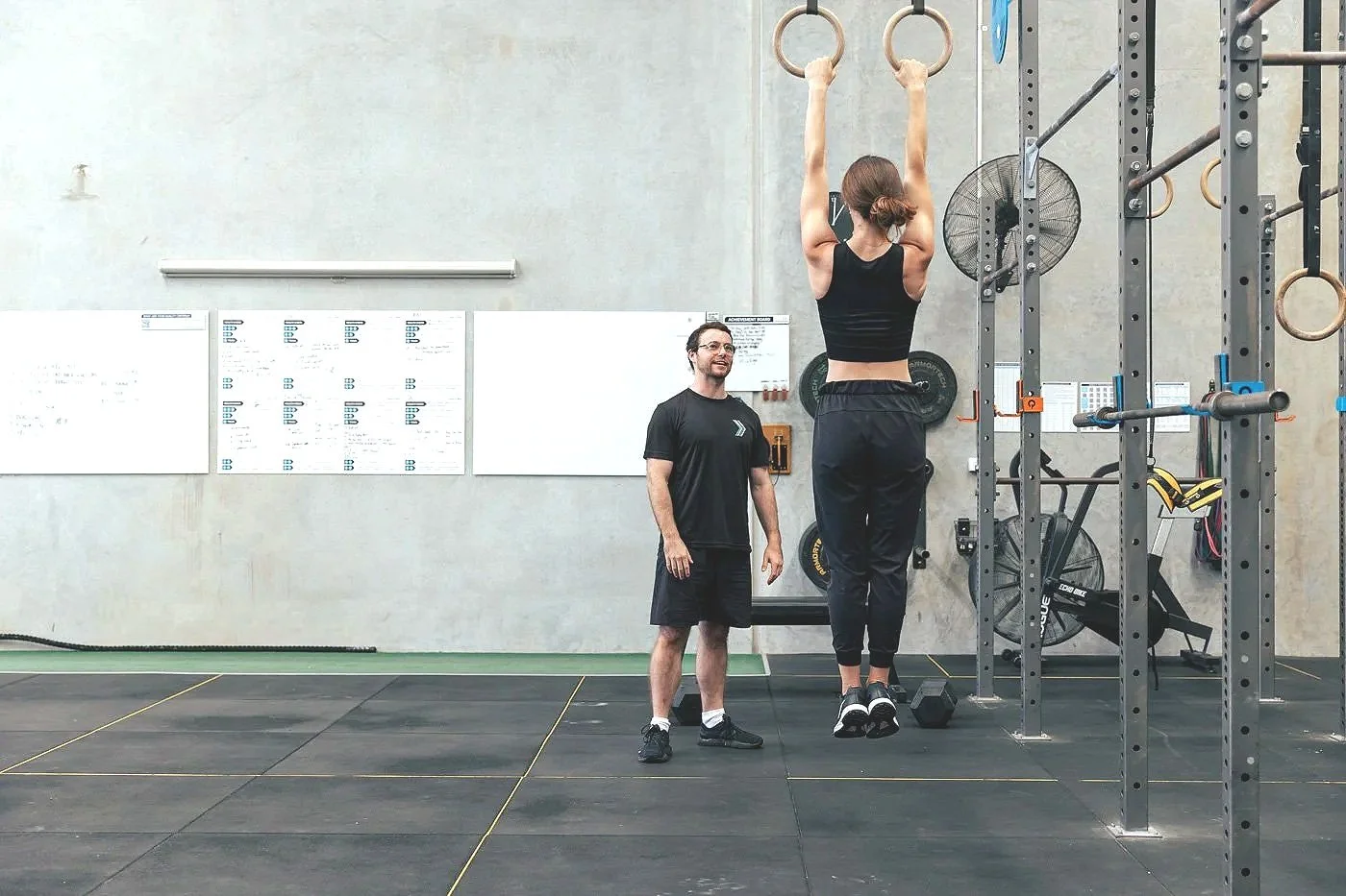 A woman with brown hair in a ponytail is hanging from gymnastic rings in a gym, assisted by a male trainer standing beside her, smiling. The gym has various equipment including pull-up bars, weights, and a black bike.