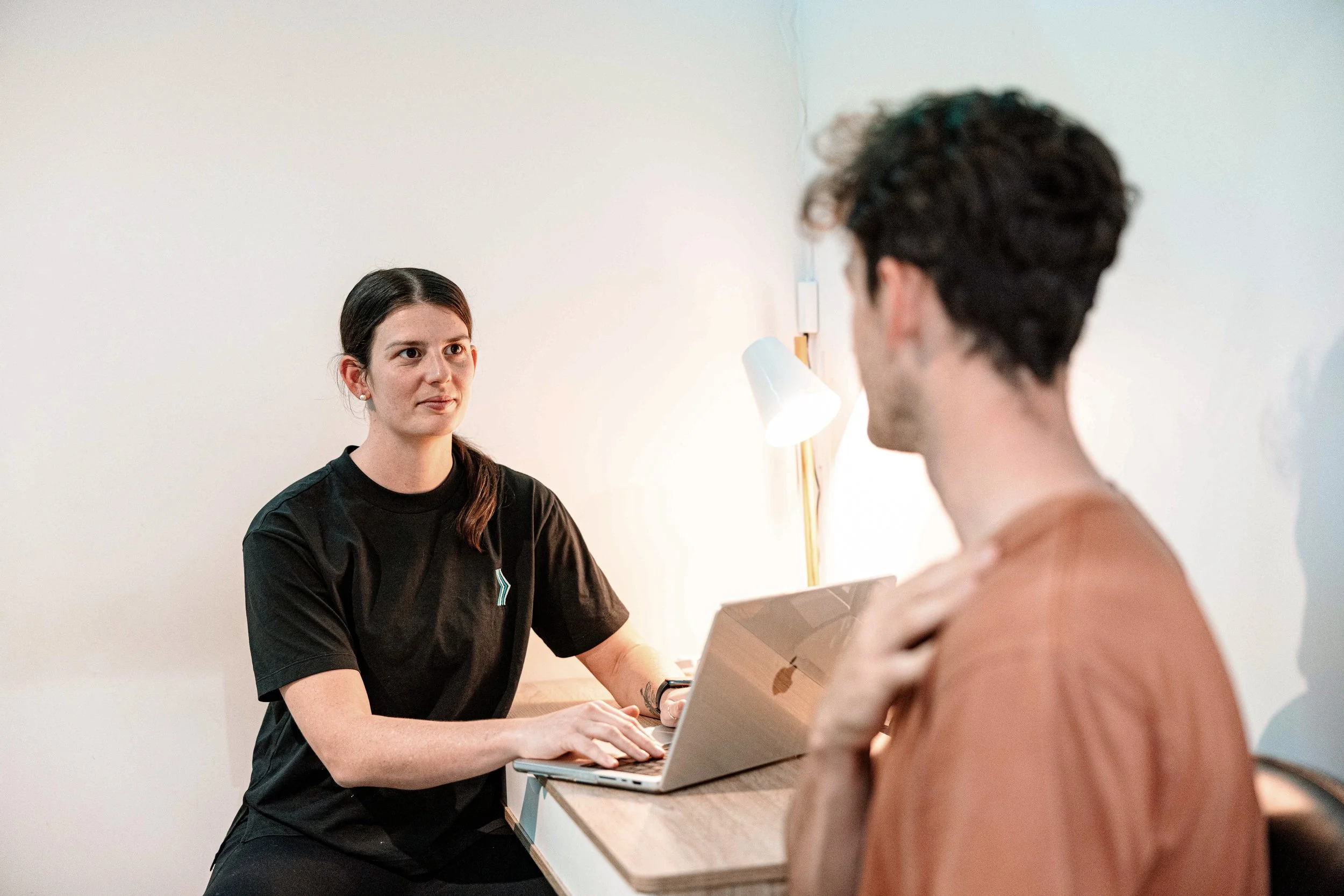 A woman in a black shirt sitting at a desk engaging in conversation with a person holding a cardboard box, in an indoor setting with white walls and a small lamp.
