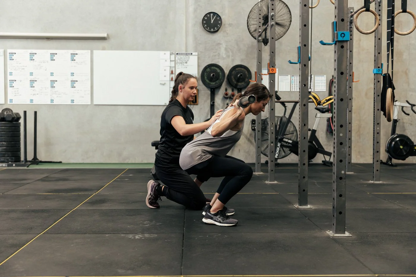 A woman is doing a squat exercise with a weight bar while a trainer supports her from behind in a gym.