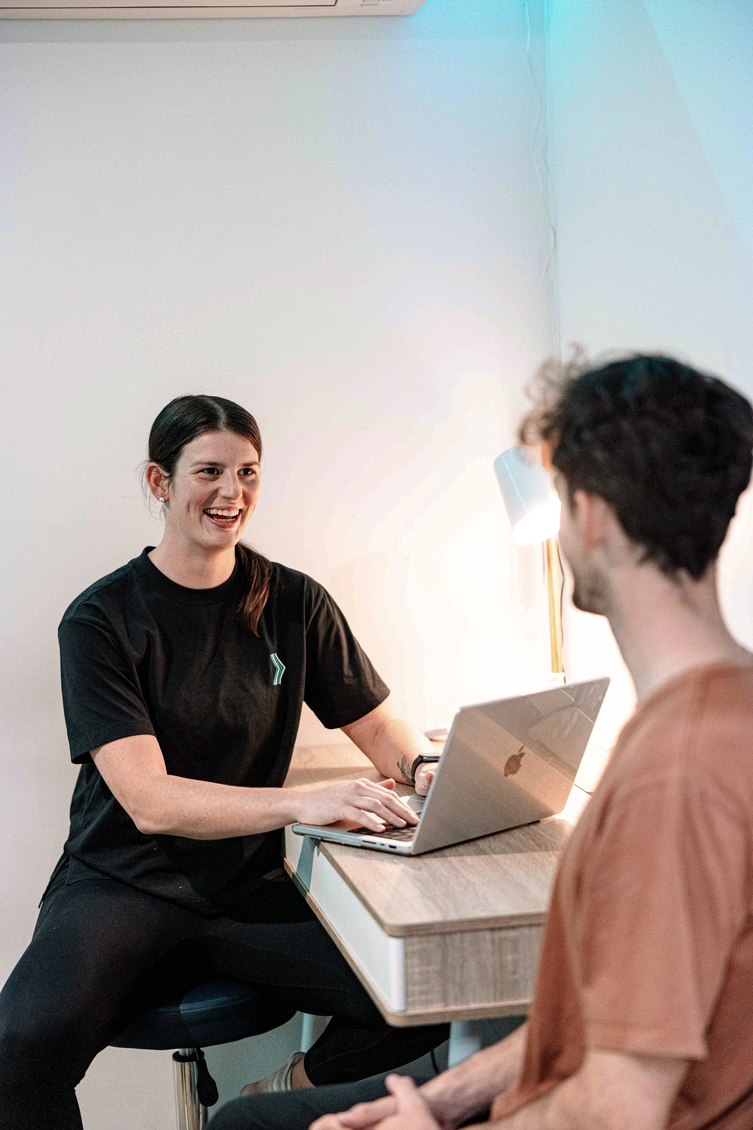 A woman with long dark hair smiling and wearing a black shirt sitting at a desk with a laptop open in front of her, talking to a person with short curly hair in a peach shirt, who is sitting across from her.