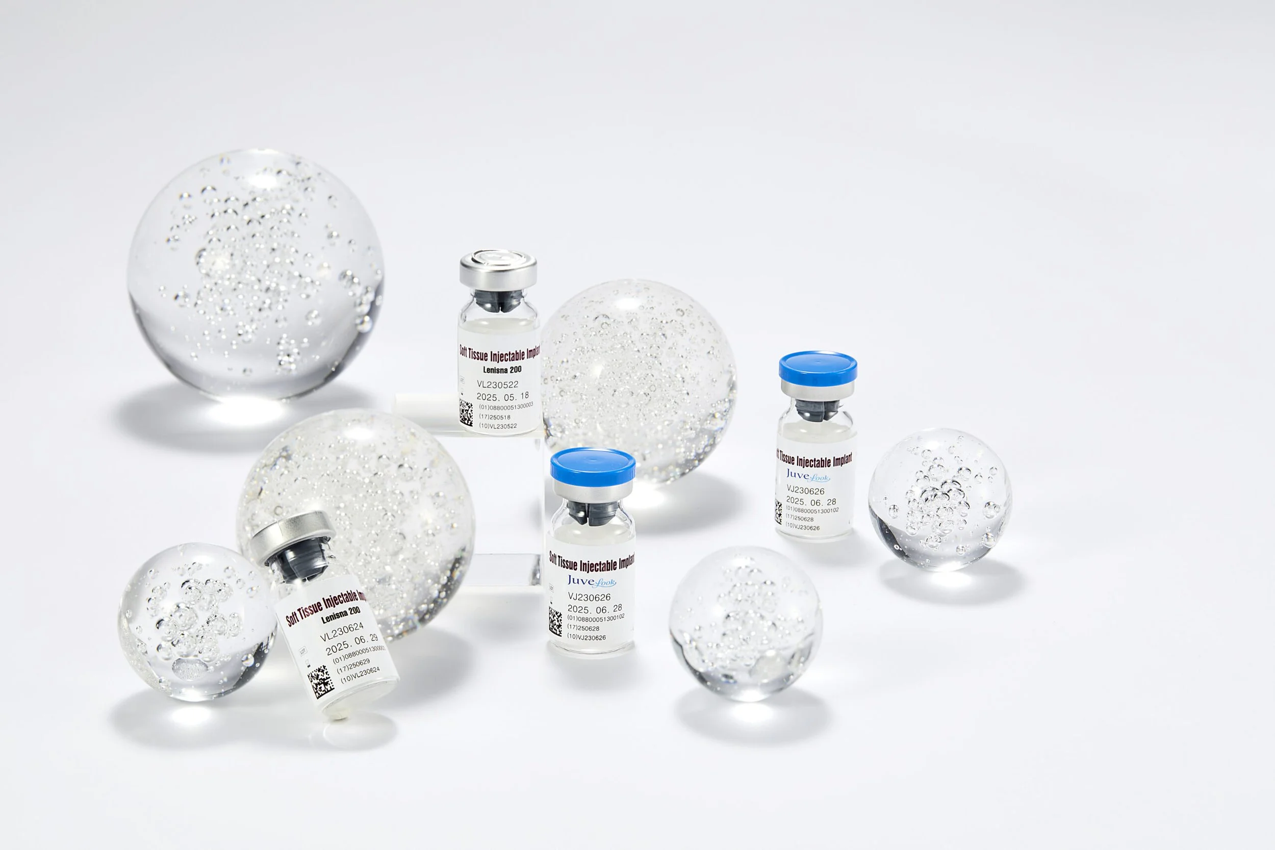 Medical vials labeled 'Soft Tissue Injectable Implant' and decorative clear glass spheres with water droplets, arranged on a white background.