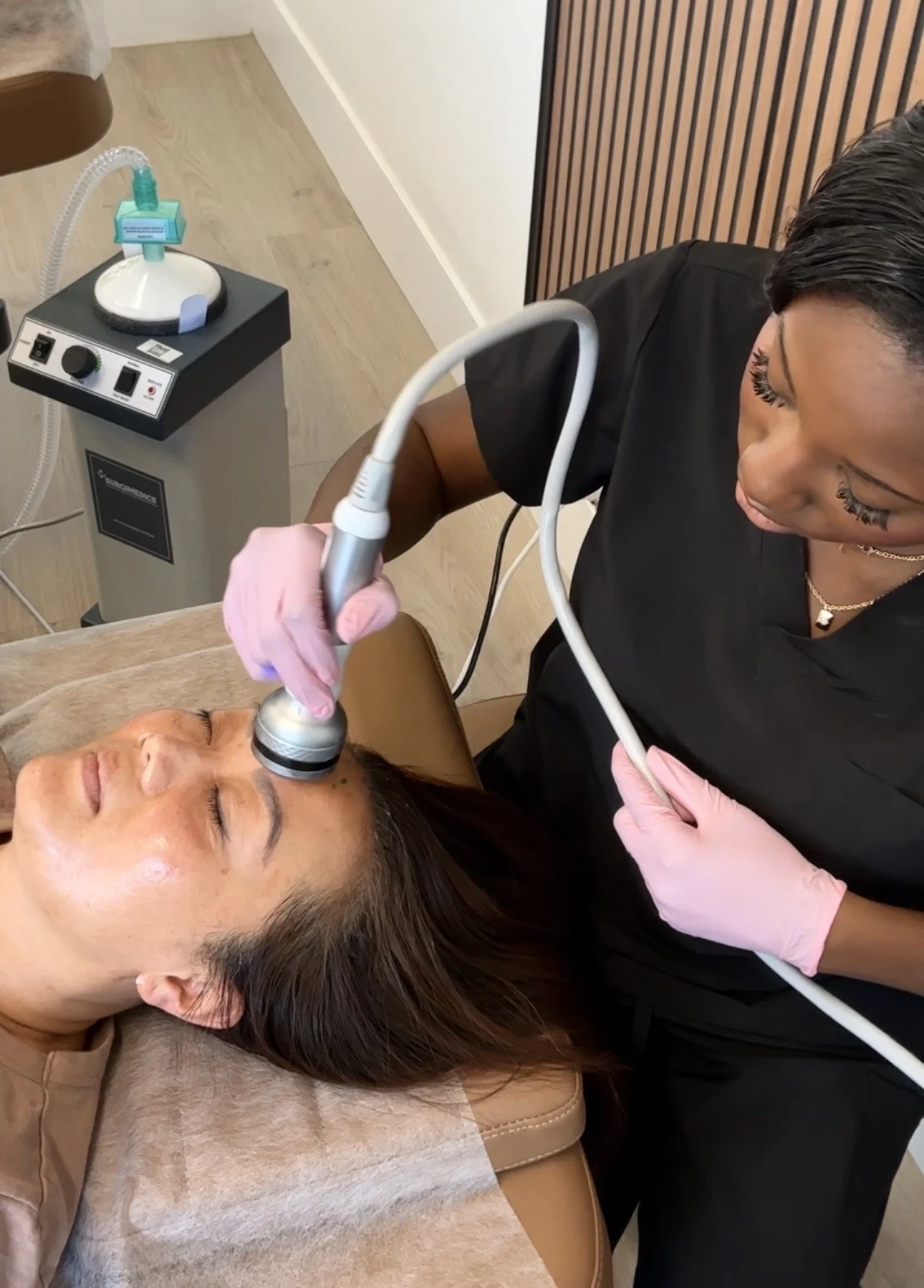 A woman receiving a facial treatment with a handheld device from a practitioner wearing pink gloves, in a skincare clinic setting.